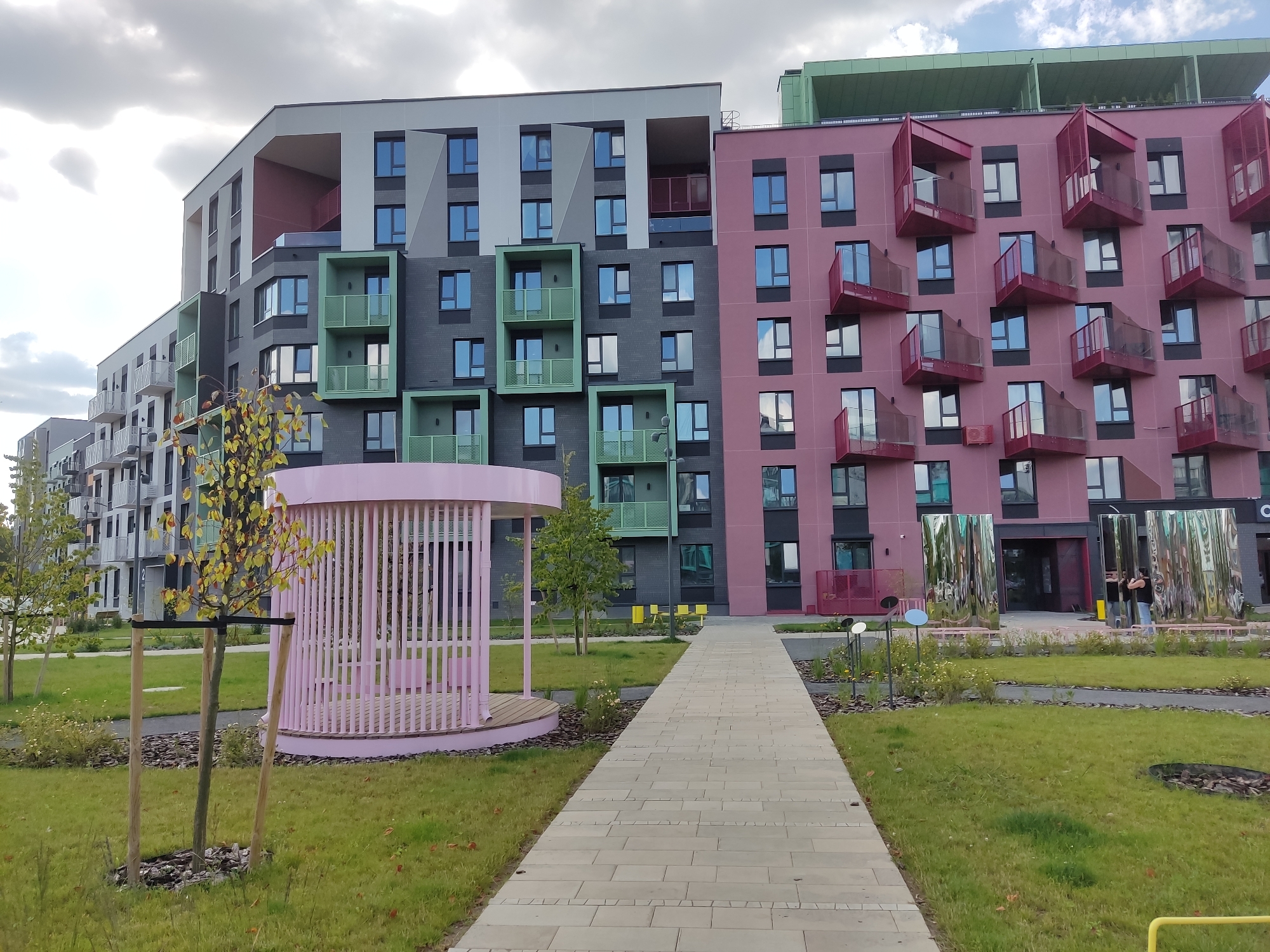 The image depicts a modern residential complex in the Baravliany rural council, Belarus. The buildings feature a colorful and geometric architectural style, with sections painted in shades of pink, dark gray, and light gray. Several balconies are adorned with bright red accents. In the foreground, a paved walkway leads towards the buildings, flanked by a grassy area with a pink, open-air gazebo or pavilion. Trees with yellowing leaves are scattered throughout the landscaping, indicating an autumn season. The sky is overcast with clouds. Two individuals are visible near the entrance of one of the buildings, interacting with reflective, sculpture-like structures.