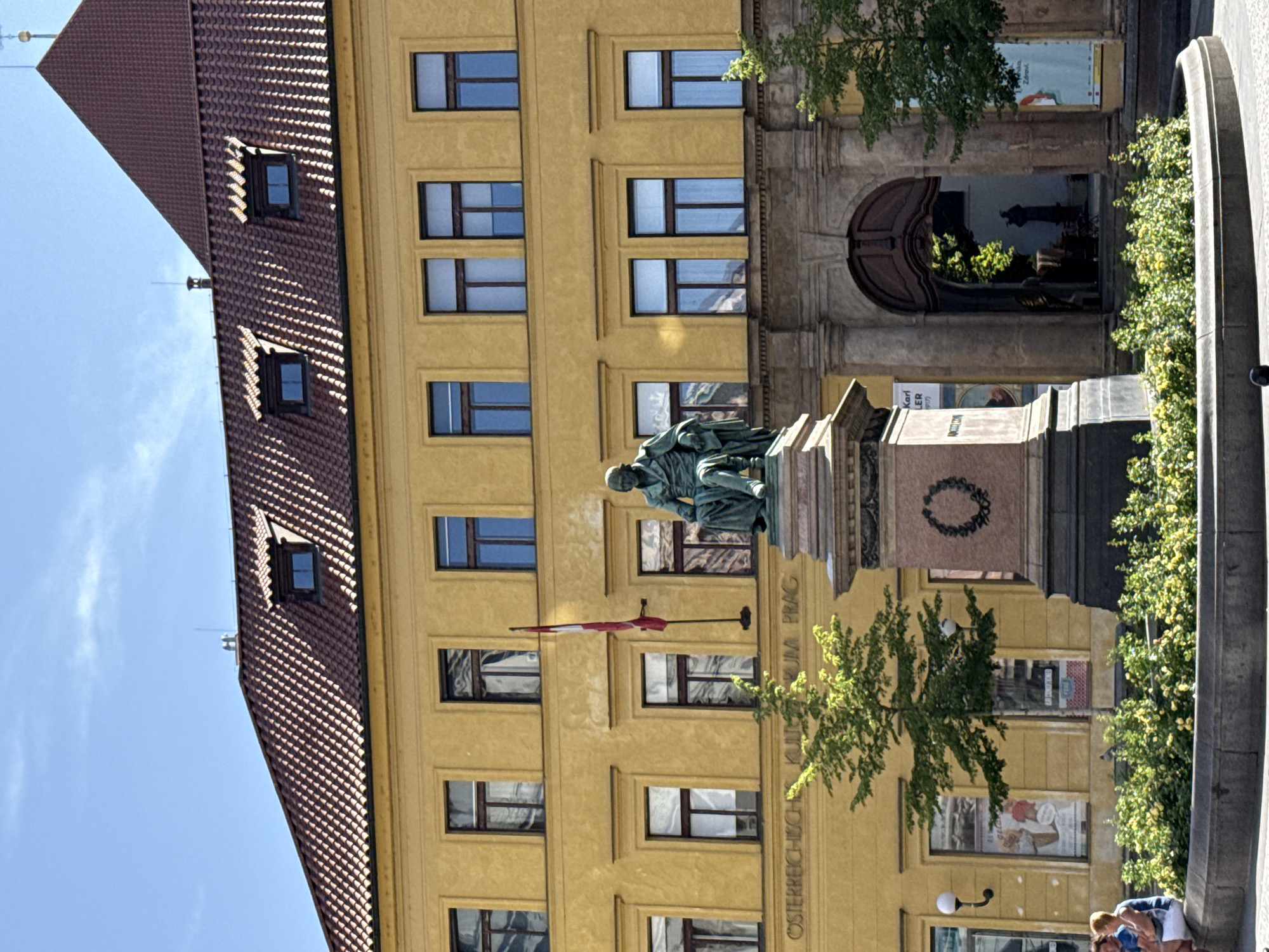 The image shows the Austrian Cultural Forum Prague, located at the Tratnovská 13. The building is a yellow, multi-story structure with a red tiled roof and dormer windows. In front of the building is a monument with a statue of a man seated in a bronze chair. The monument is surrounded by a circular planter filled with green bushes and flowering plants. Two people are visible sitting on the edge of the planter on the left side of the image. There is a flag hanging on the building to the left of the monument, and an arched entryway is visible to the right. Trees with green foliage are present in front of the building and to the right of the monument. The sky above is blue with some clouds.