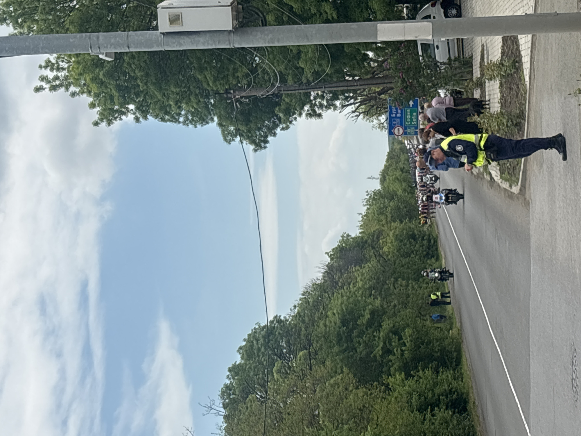 The image depicts a road in Burgas, Bulgaria, lined with green trees and a clear blue sky with white clouds. A police officer wearing a yellow vest and dark uniform stands on the right side of the road, near a group of people watching a procession of motorcycles and cyclists heading along the road. A road sign indicating directions to Burgas and Sofia is visible near the crowd. A white van is parked further back along the road. Overhead power lines and utility poles are visible.