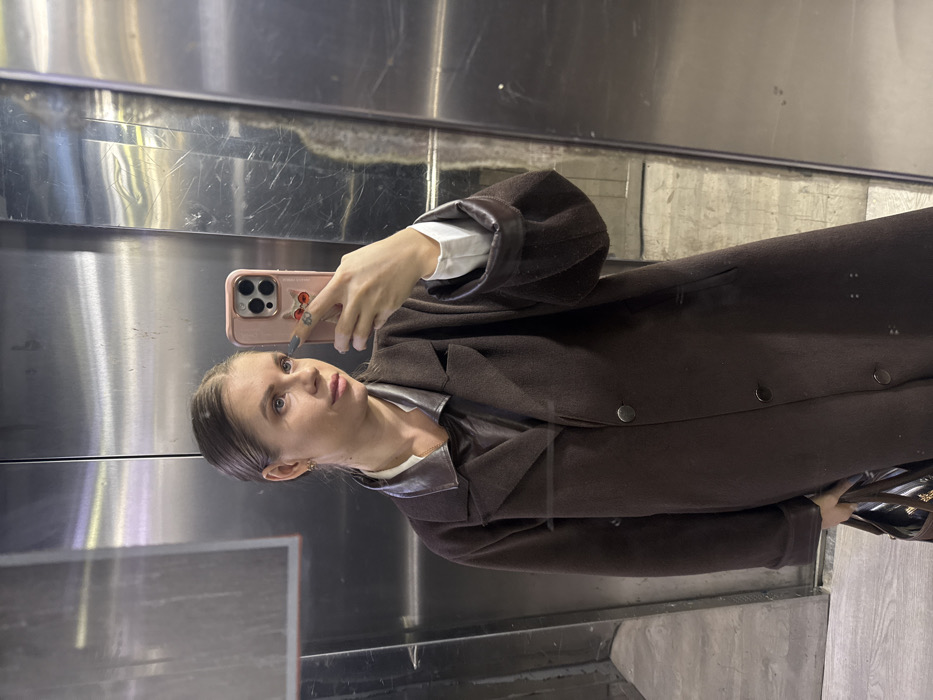 A woman takes a selfie in a mirrored elevator. She wears a long, dark brown coat with leather accents, a white shirt, and gold earrings. She holds a pink phone with a cat case. The elevator walls are mirrored and show smudges and wear. The floor is a mottled gray.