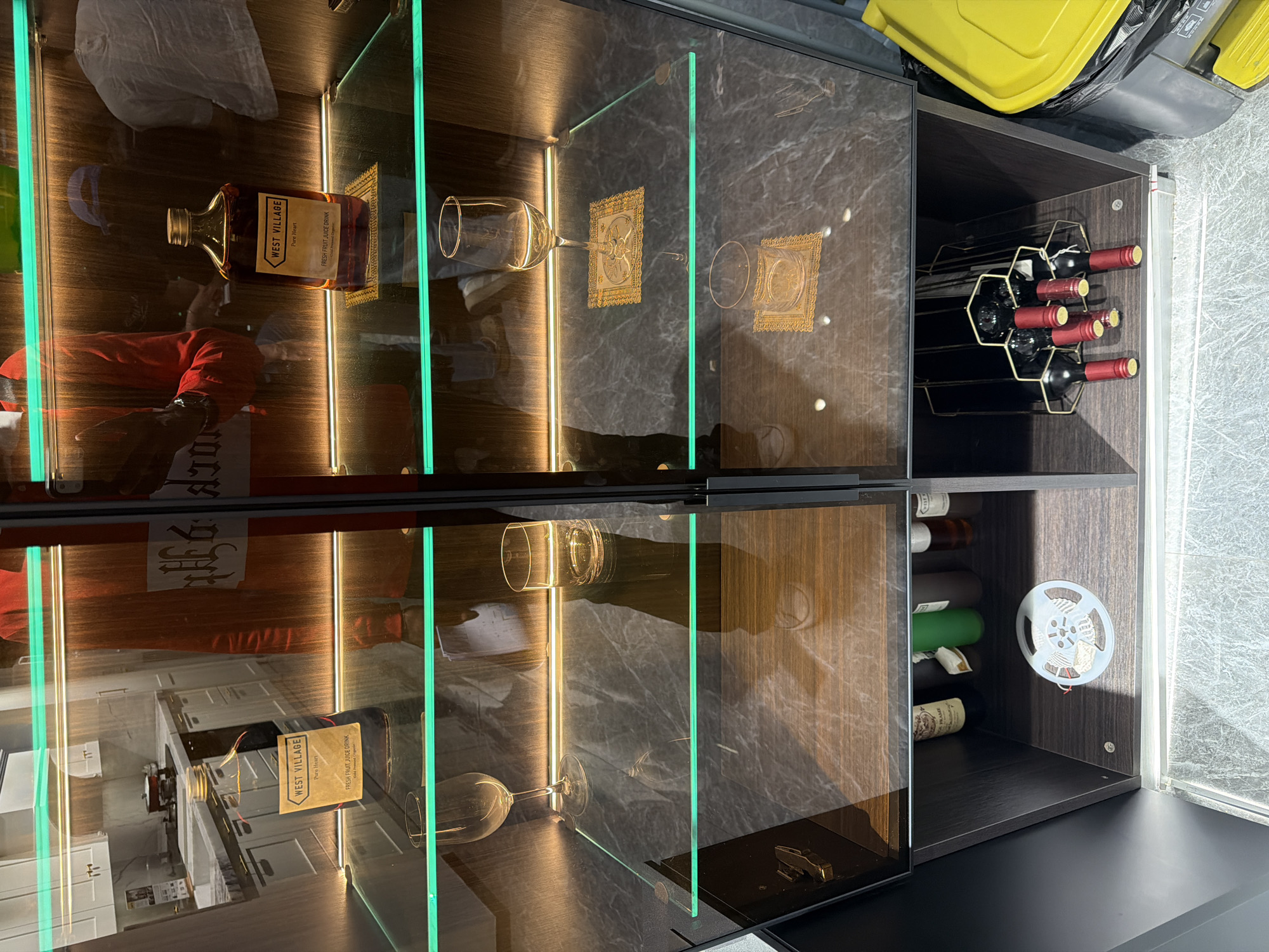 The image displays a modern, illuminated display cabinet, appearing to be rotated 90 degrees clockwise from its natural upright position. The cabinet features multiple glass shelves, framed by integrated LED lighting strips that emit both warm amber and greenish hues, creating a sophisticated ambiance. The structure combines dark wood or laminate panels with a marble-like pattern.
On the upper left shelf, an amber-colored glass bottle is prominently displayed, featuring a label that reads "WEST VILLAGE," "Pure Heart," and "FRESH FRUIT JUICE DRINK." Reflections on the glass front show parts of an indoor environment, including what appears to be a person wearing a red top. Beside the bottle, an empty wine glass lies on its side, near a decorative gold-colored square item. The upper right section of the cabinet houses a wine rack, holding several dark wine bottles with distinct red and gold foil caps.
The lower section of the cabinet mirrors the upper left, with another "WEST VILLAGE Pure Heart FRESH FRUIT JUICE DRINK" bottle, along with more reflections of an interior space, potentially a kitchen with counters and cabinets. An empty glass is also visible on its side on a lower shelf. On the right side of the lower cabinet, a white spool of material, possibly LED strip or wiring, is mounted on the inner wall.
Above the cabinet (which would be to the right if the image were correctly oriented), a bright yellow plastic object, possibly part of a container, and a dark trash bag are visible. The surrounding floor area appears to be textured or tiled. The visible text elements include "WEST VILLAGE," "Pure Heart," "FRESH FRUIT JUICE DRINK," and the word "FRAME" which appears as a reflection or part of the cabinet's design. The scene provides no specific time of day or emotional cues. The location context is Chancheng District, China, though no direct visual cues in the image confirm this.