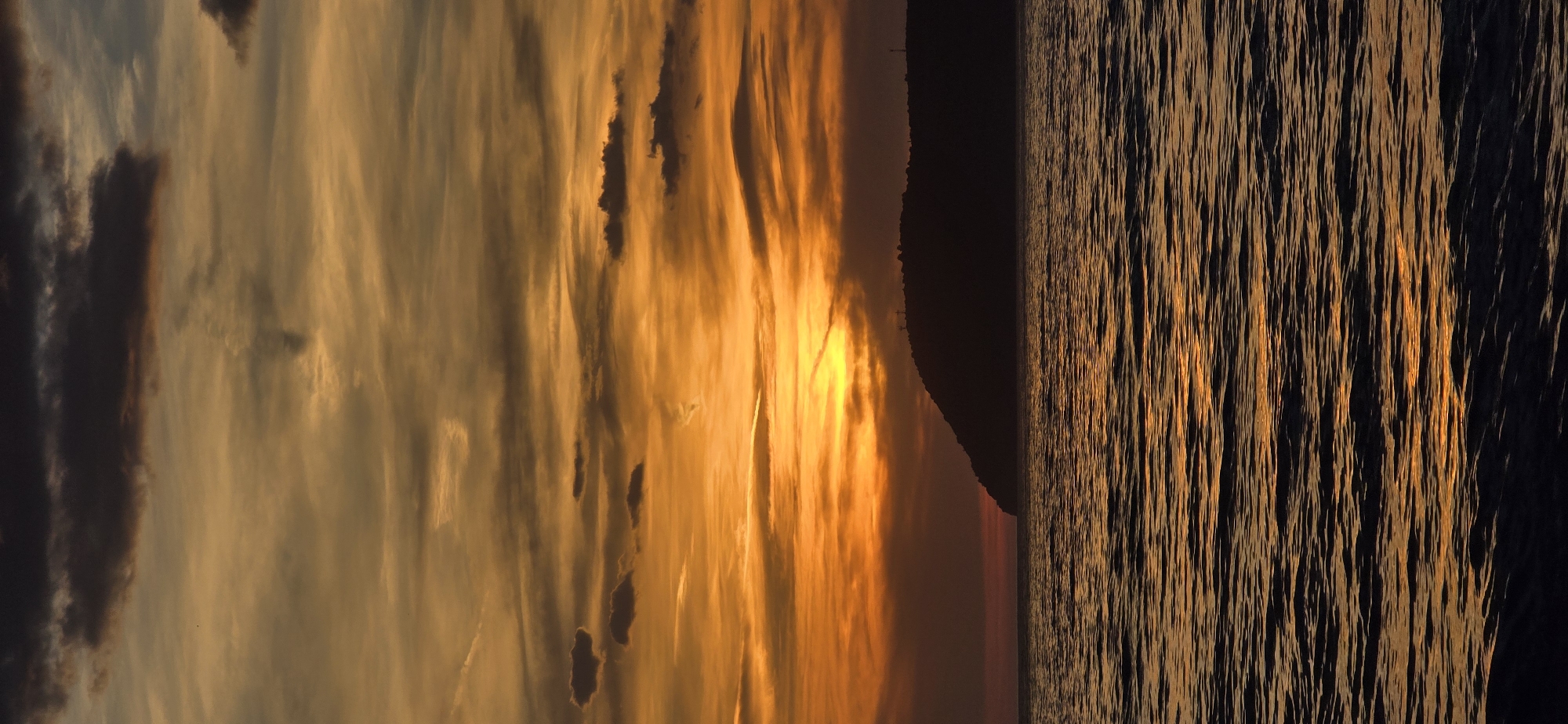 The image depicts a sunset scene in Bar, Montenegro. The left side shows a sky with layered, streaky clouds ranging in color from dark gray to golden orange, illuminated by the setting sun. The sun itself appears as a bright, diffused glow behind the clouds.
The right side of the image shows water with small, reflective waves. The water's color ranges from dark to shimmering orange where it reflects the sunlight.
A dark, silhouetted landmass is visible between the sky and the water, indicating a coastline.