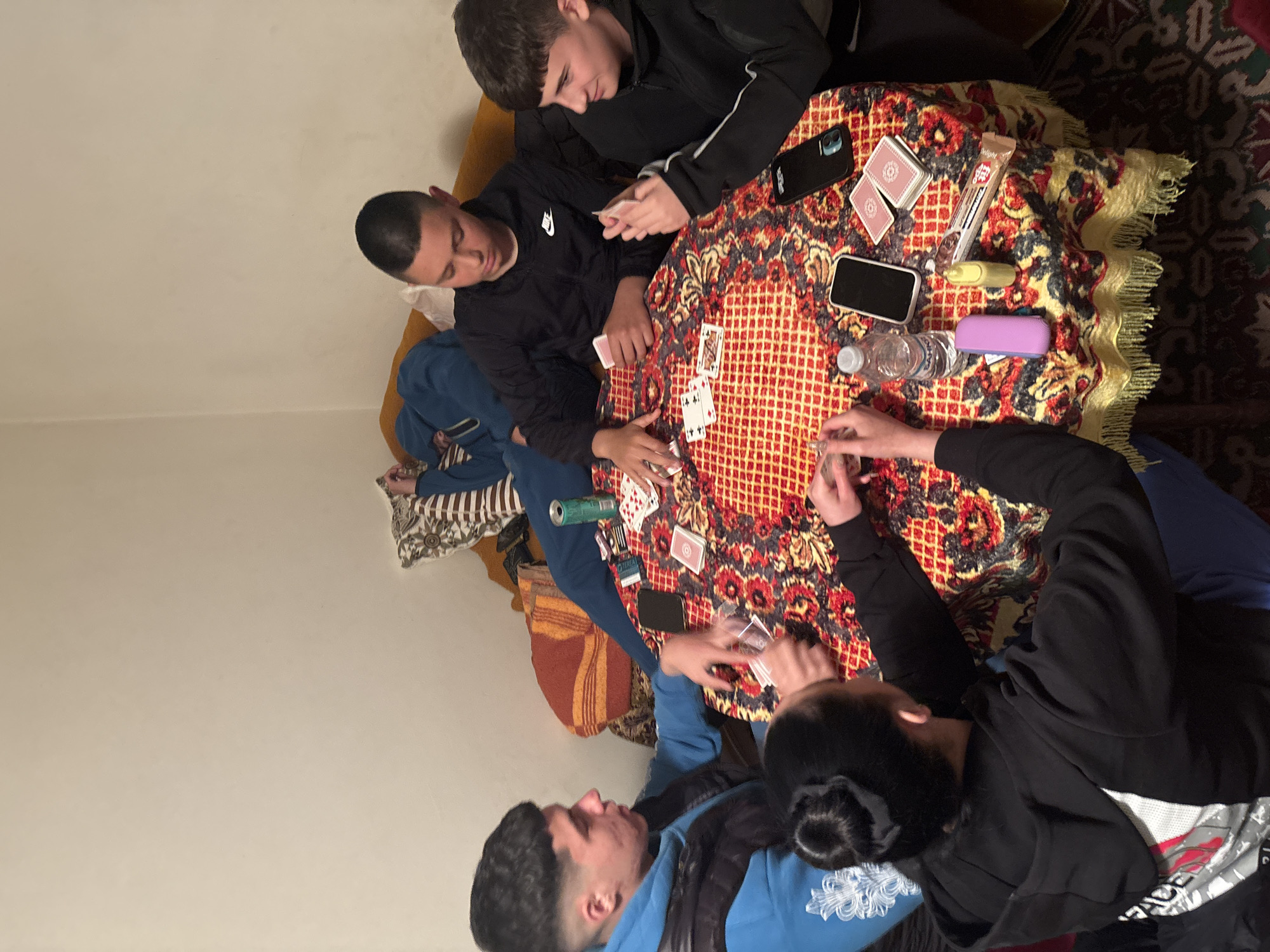 Four young men are playing cards around a round table covered with an ornate tablecloth, indoor. The table also contains two smartphones, a water bottle, snack bar, and other miscellaneous items. The men are casually dressed in hoodies and sweatshirts. They are seated on a variety of cushions and blankets. The location of the image is Goritsa, Bulgaria.