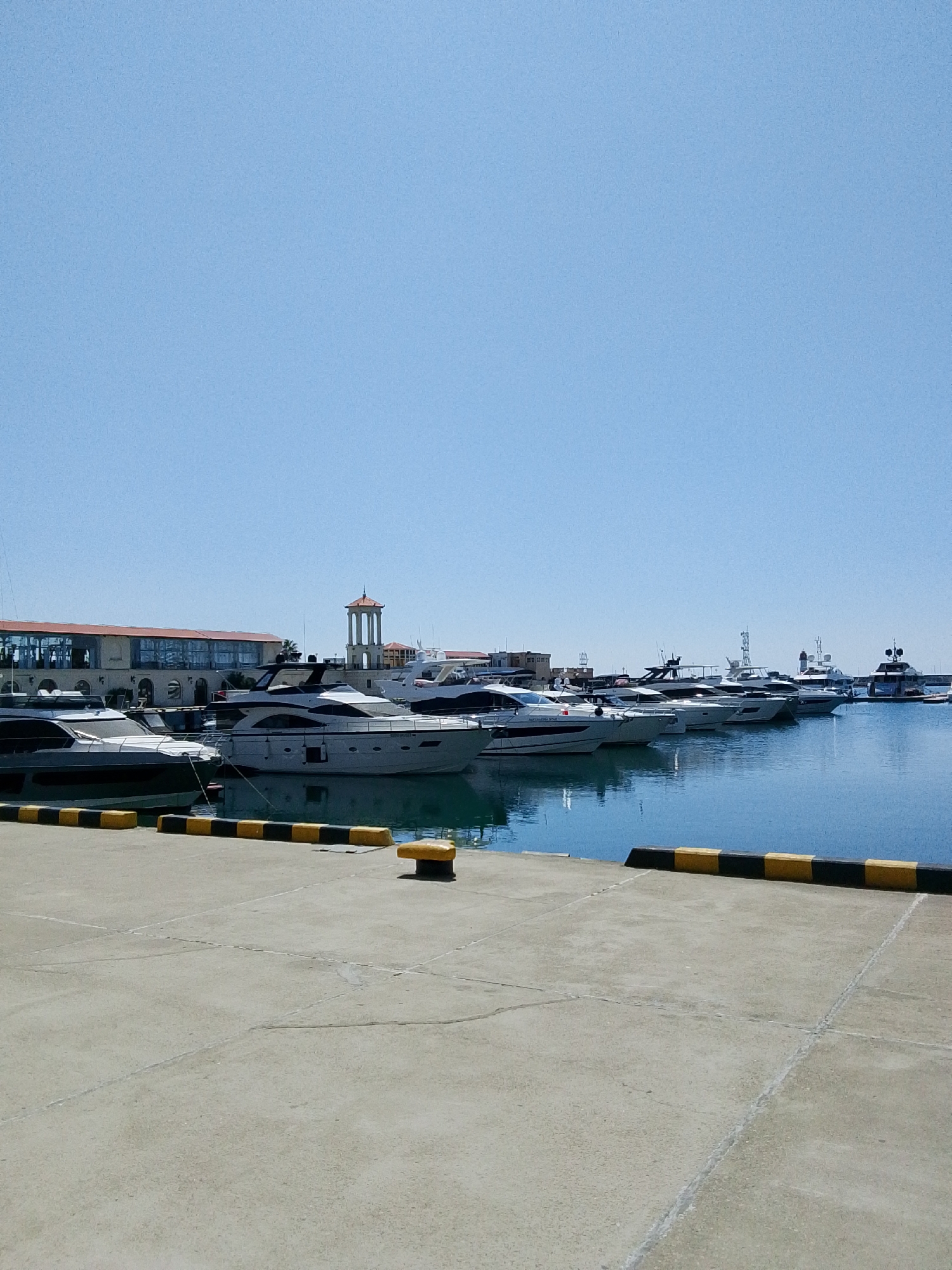 The image shows a bright, sunny day at a marina. The sky is clear and blue, and the water is a deep blue. In the foreground, a concrete pier is visible, with yellow and black bollards along the edge. Several yachts and boats are docked in the background. Some of the yachts are large and luxurious, while others are smaller. A building with an orange roof and large windows is visible on the left side of the image. A tall, tower-like structure with a red roof is also present in the background. The scene appears peaceful and serene, with no people visible in the image. The overall impression is one of a well-maintained and affluent marina on a beautiful day.