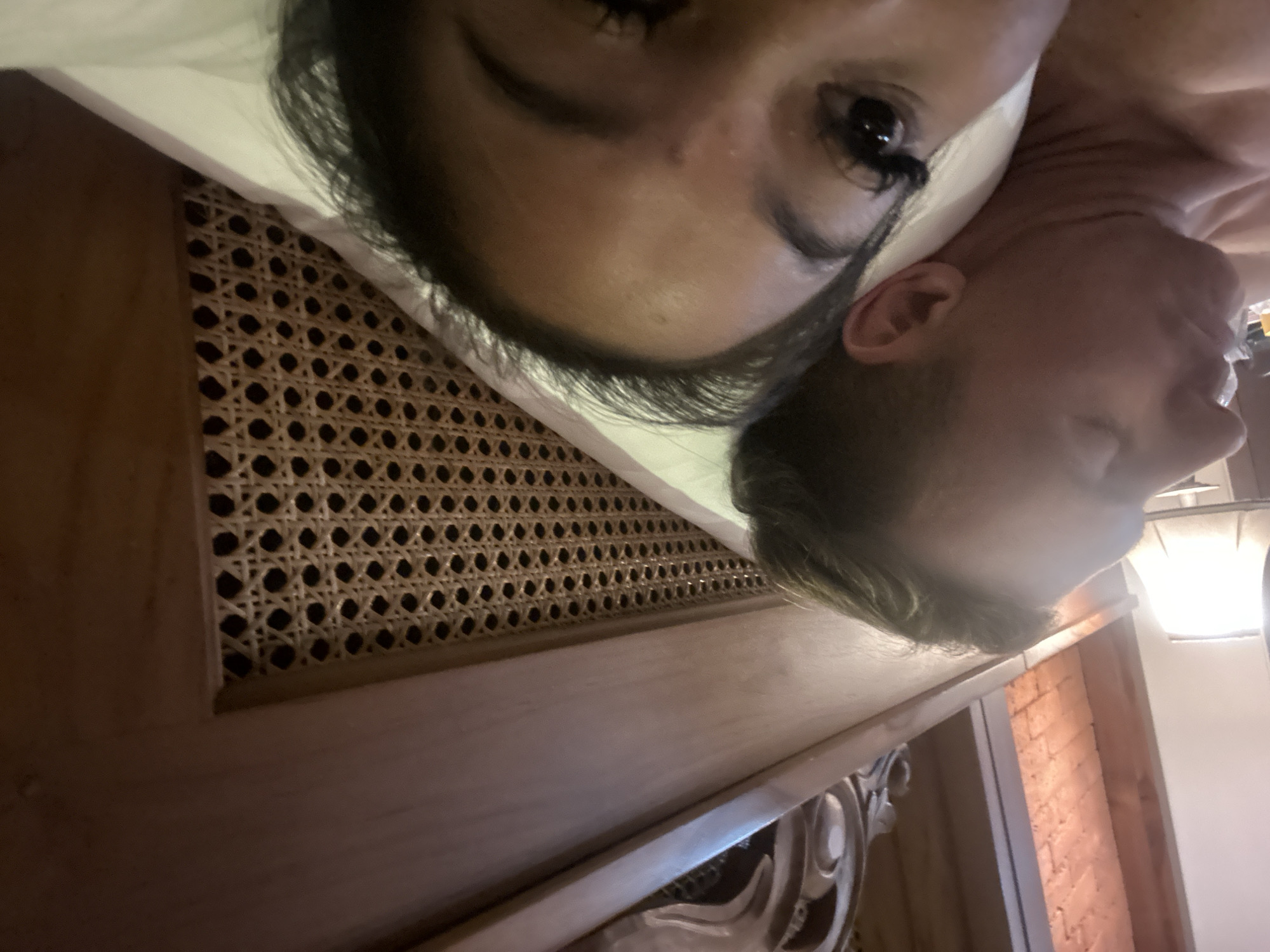 A selfie-style, low-angle shot depicts two individuals lying down indoors. A female with dark hair and eyelashes is positioned closer to the camera, next to a male with light-colored hair and a beard. The background features a wooden panel with a cane webbing design, and a section of what appears to be a brick wall. Based on the provided location, it is possible this scene is occurring in a building within the Ubud District of Indonesia.