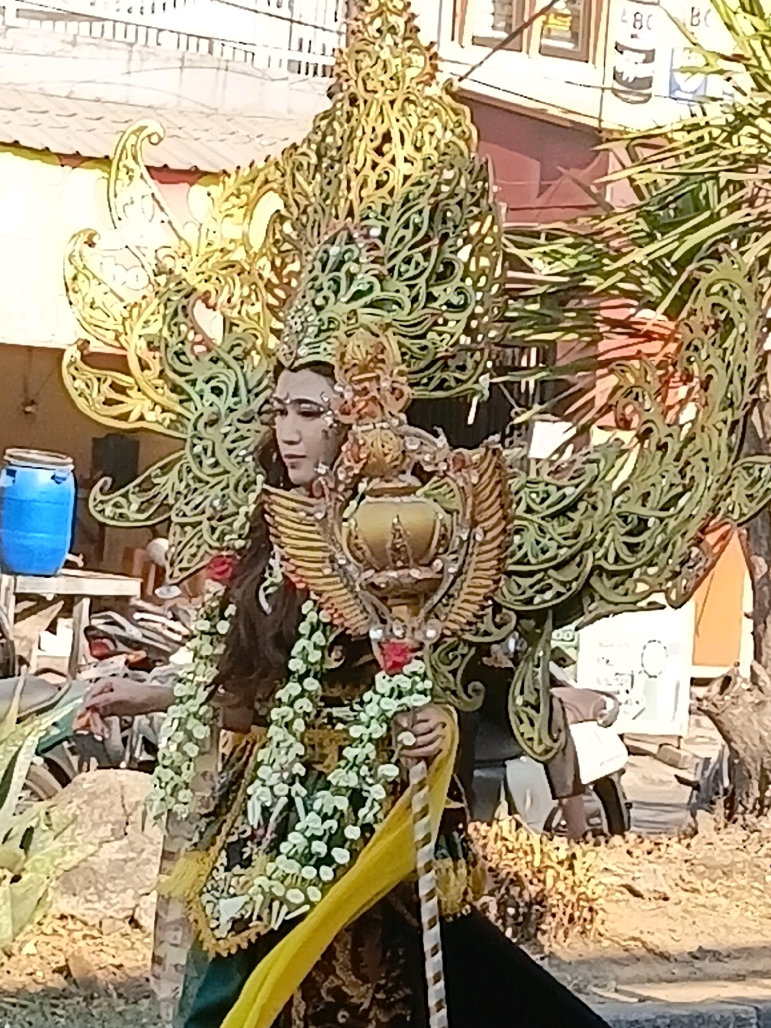 In Wadungasri, Indonesia, a person stands adorned in an elaborate, traditional costume. The costume features a large, intricate headpiece resembling wings or a headdress, with ornate, green, scroll-like designs. They wear a collar made of white flowers and a golden staff with a crown-like embellishment. The person's face is painted with dramatic makeup, and they have long, dark hair. Behind them, a blue barrel sits on a wooden surface, and a building with windows and signage is visible. The ground is covered in dry leaves and debris. The scene appears to be outdoors, possibly during an event or festival.