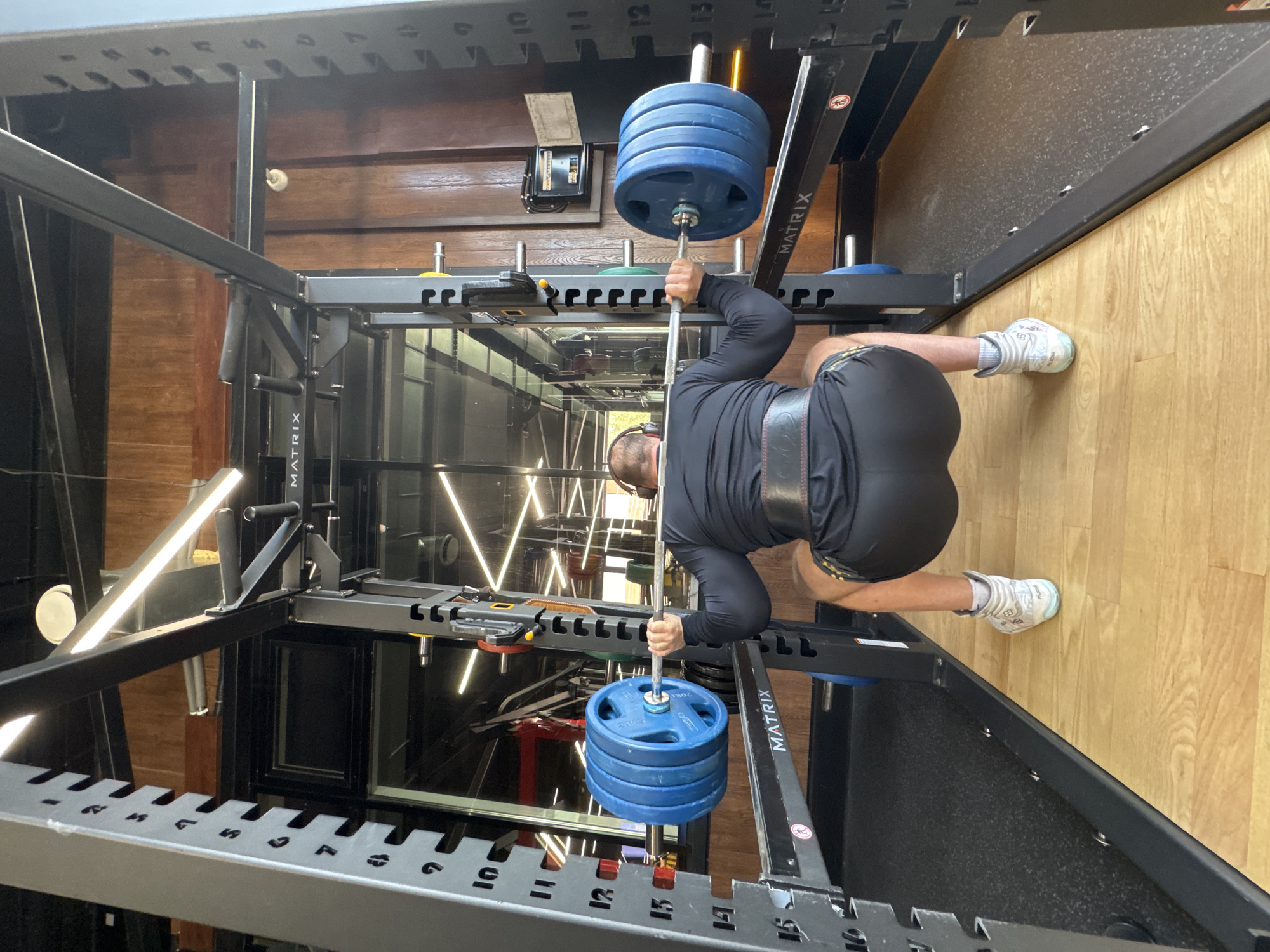 The image, when viewed in its correct upright orientation, captures a male individual performing weight training inside a modern indoor gym. The man, clad in a long-sleeved black athletic shirt, black shorts, a weightlifting belt, and light-colored athletic shoes, is gripping a barbell loaded with four blue weight plates on each side. He is positioned within a black Matrix-branded squat rack or smith machine, with the barbell resting on or being lifted from a lower safety pin or J-cup. His posture suggests he is either performing a deadlift, a bent-over row, or the lower phase of a squat.
The gym features light wooden flooring and dark wooden paneling on the walls. Reflections of bright, geometric LED lights are visible in the background, suggesting glass surfaces or mirrors. The equipment itself is sturdy, with numerical markings along its frame (e.g., numbers 10 through 17 on the lower rail, and letters like S, P, F, R, G on the upper rail), indicating different height adjustments. The weight plates are also branded "MATRIX." The man wears headphones or earbuds and appears concentrated on his workout. The setting is well-lit, likely by a combination of natural and artificial light.