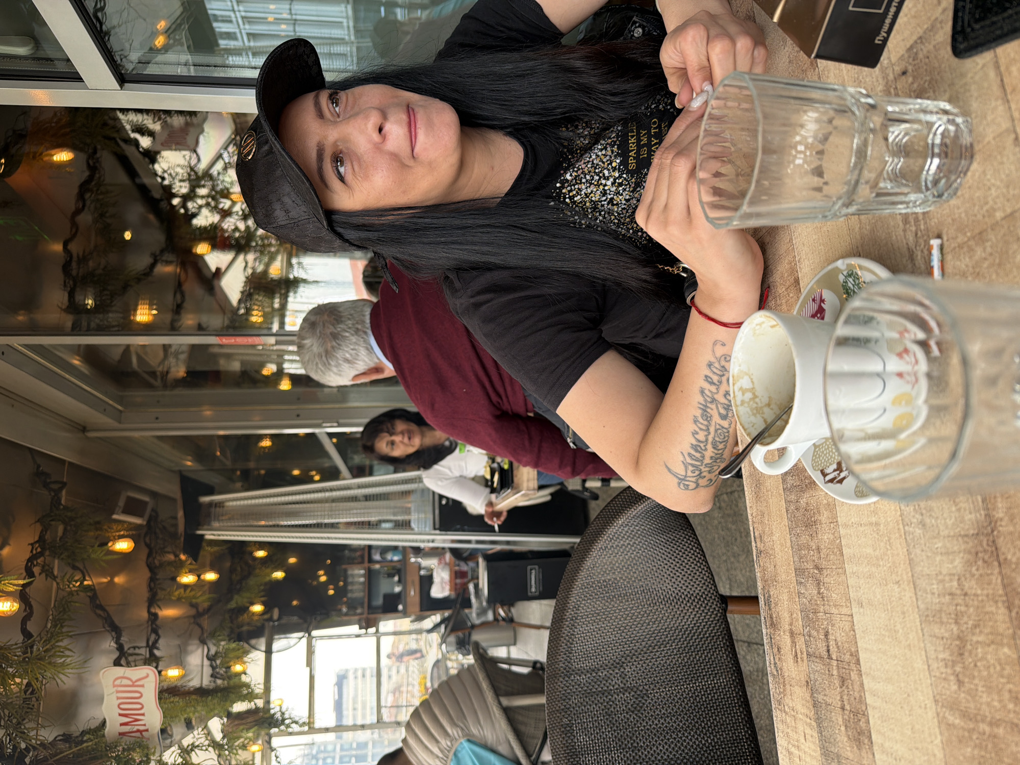 The image depicts a woman wearing a black t-shirt, black cap, and long black hair, seated at a wooden table at an establishment in Sofia, Bulgaria. An empty glass and a used coffee cup sit on the table. The woman has a tattoo on her left arm and is wearing a red bracelet. Behind her, there are other people, including a person with a white shirt and black hair, and another person with a grey hair and maroon sweater. Decorative lights and vegetation are visible in the background, along with architectural elements suggesting an enclosed or semi-enclosed space such as a patio or restaurant with a view of the cityscape. A sign with the word "AMOUR" is present among the decorations.