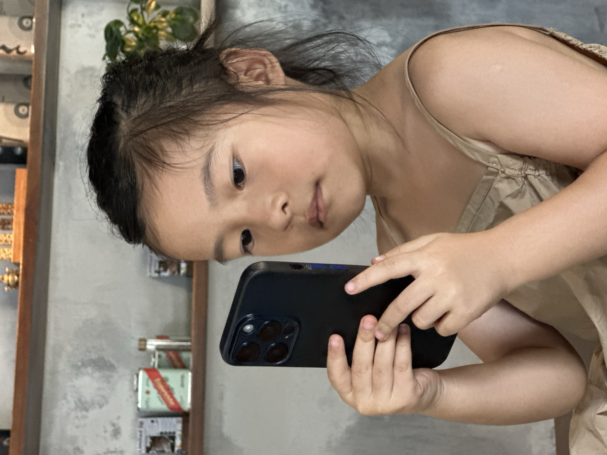 A young East Asian girl, with dark hair pulled back in a bun and wispy strands framing her face, is shown in a close-up shot. She is wearing a beige tank top and holding a black smartphone horizontally in her hands, seemingly engrossed in its screen. Her gaze is directed downwards towards the device. The background suggests an indoor setting with a textured grey wall and a wooden shelf above, displaying various items like tins and possibly jars. The lighting is natural, casting soft shadows. The scene is static, capturing a moment of solitary engagement with technology. The image does not provide any specific visual cues that would allow for the localization of this scene within Đà Nẵng, Vietnam.