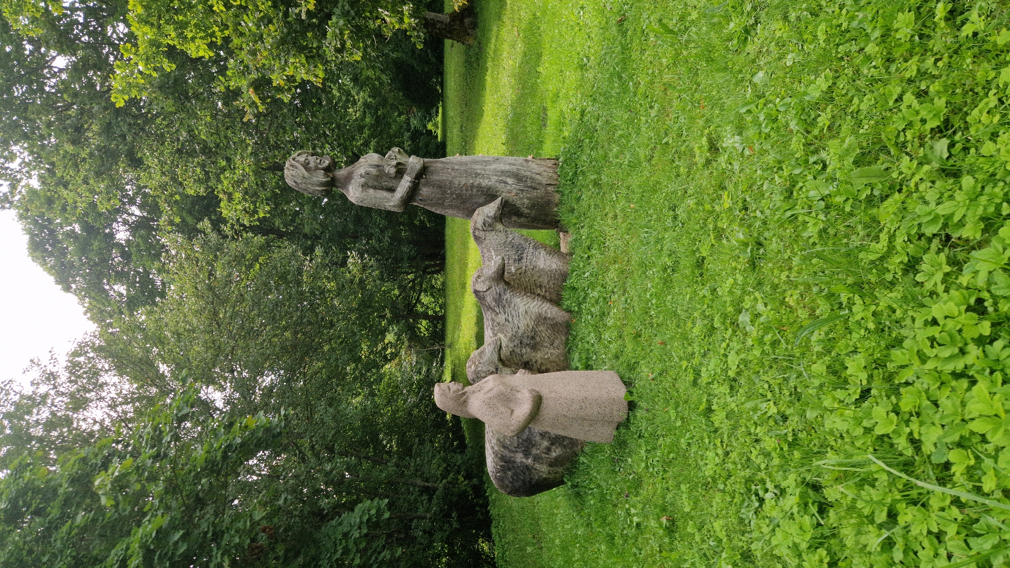 This is a photograph taken in the Kazdanga municipality of Latvia. In the foreground, lush green grass covers the ground. Two stone sculptures are visible: on the left, a female figure in a long dress stands facing right, and to her right, a series of three carved wooden sheep are arranged in a line. Behind the sheep, a tall, slender wooden sculpture of a female figure holding a bouquet stands facing left. The background is dominated by a dense forest of green trees under a bright sky.
