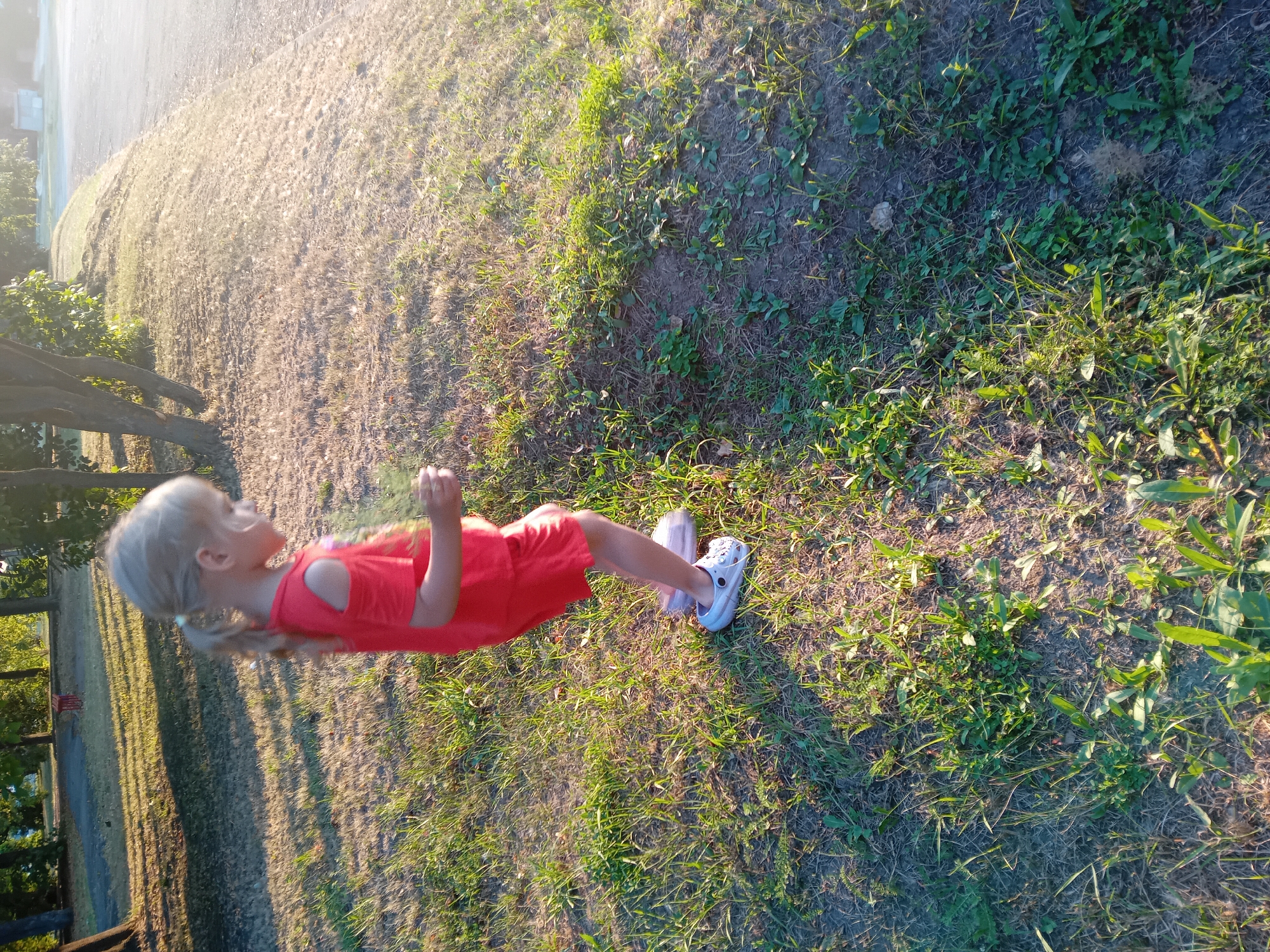 In Chuhuiv, Ukraine, a young girl with blonde hair in a ponytail, wearing a red off-the-shoulder dress and white clogs, stands on a grassy embankment. The sun casts long shadows from trees onto the grass and a paved area to the right of the embankment. The girl is holding a small green object in her right hand. The environment appears to be a park or recreational area.