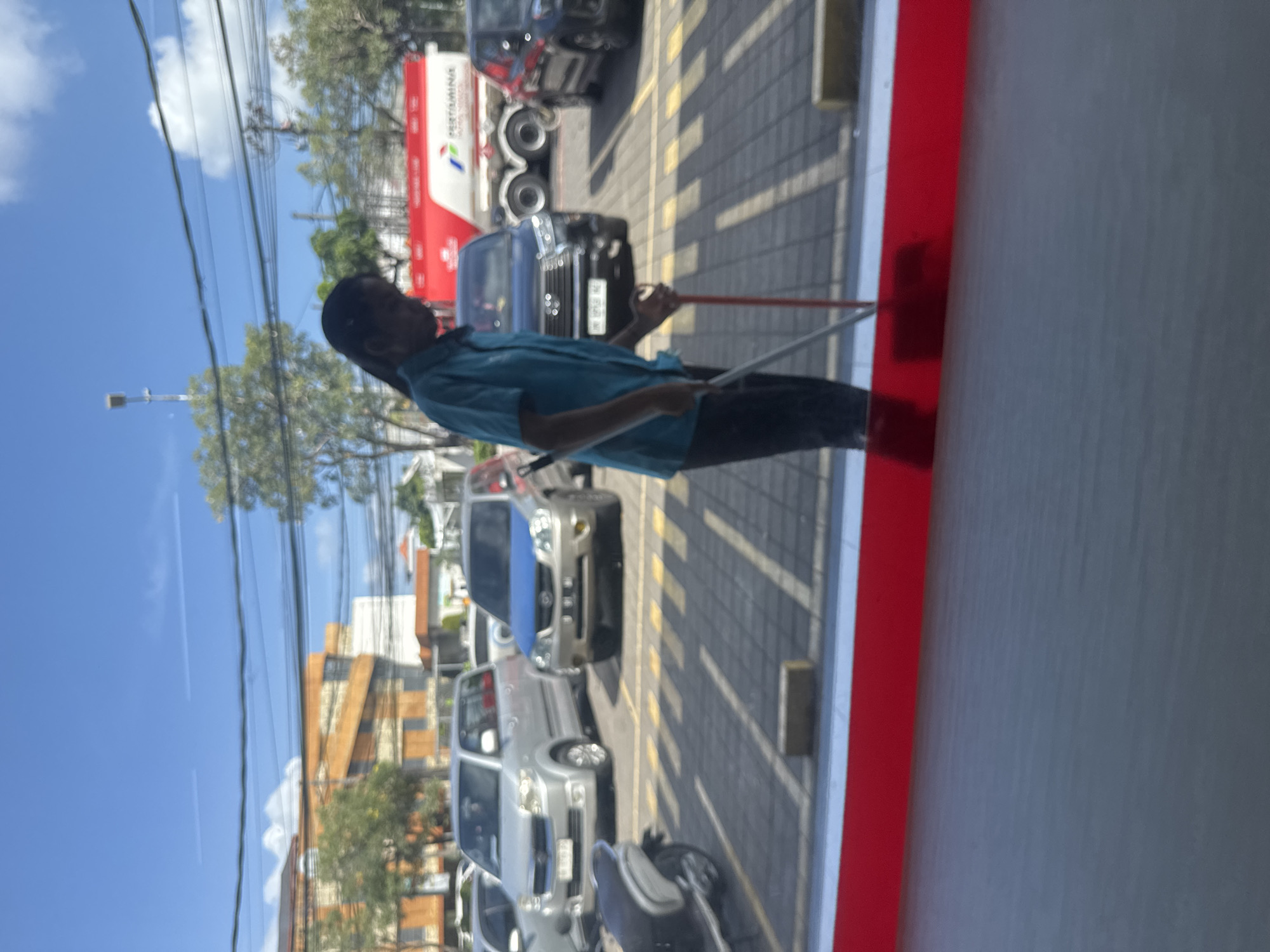 A woman in a teal uniform is sweeping a parking lot in Kuta, Indonesia. She is holding a broom with a red handle and is sweeping the pavement. In the background, there are several cars parked in the lot, including a silver minivan and a black SUV. A large red and white Pertamina fuel truck is visible in the distance. The sky is blue with some white clouds. The scene appears to be a sunny day.