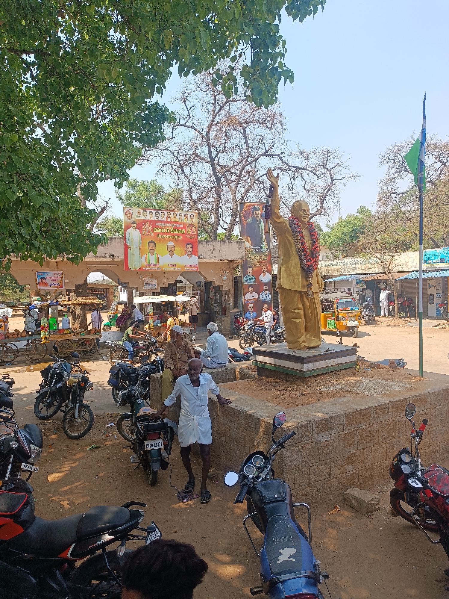 The image shows a public square in Chilamathur, India. A golden statue of a man wearing a garland stands on a raised platform. Political banners hang above an arched structure in the background. Several people are in the square, some sitting and others standing near parked motorcycles. A man in traditional clothing stands beside a motorcycle. A flag flutters beside the statue. The ground is dusty and unpaved. Trees are visible, casting shadows on the scene. Several motorcycles and auto rickshaws are parked around the square. Small shops line the street in the background.