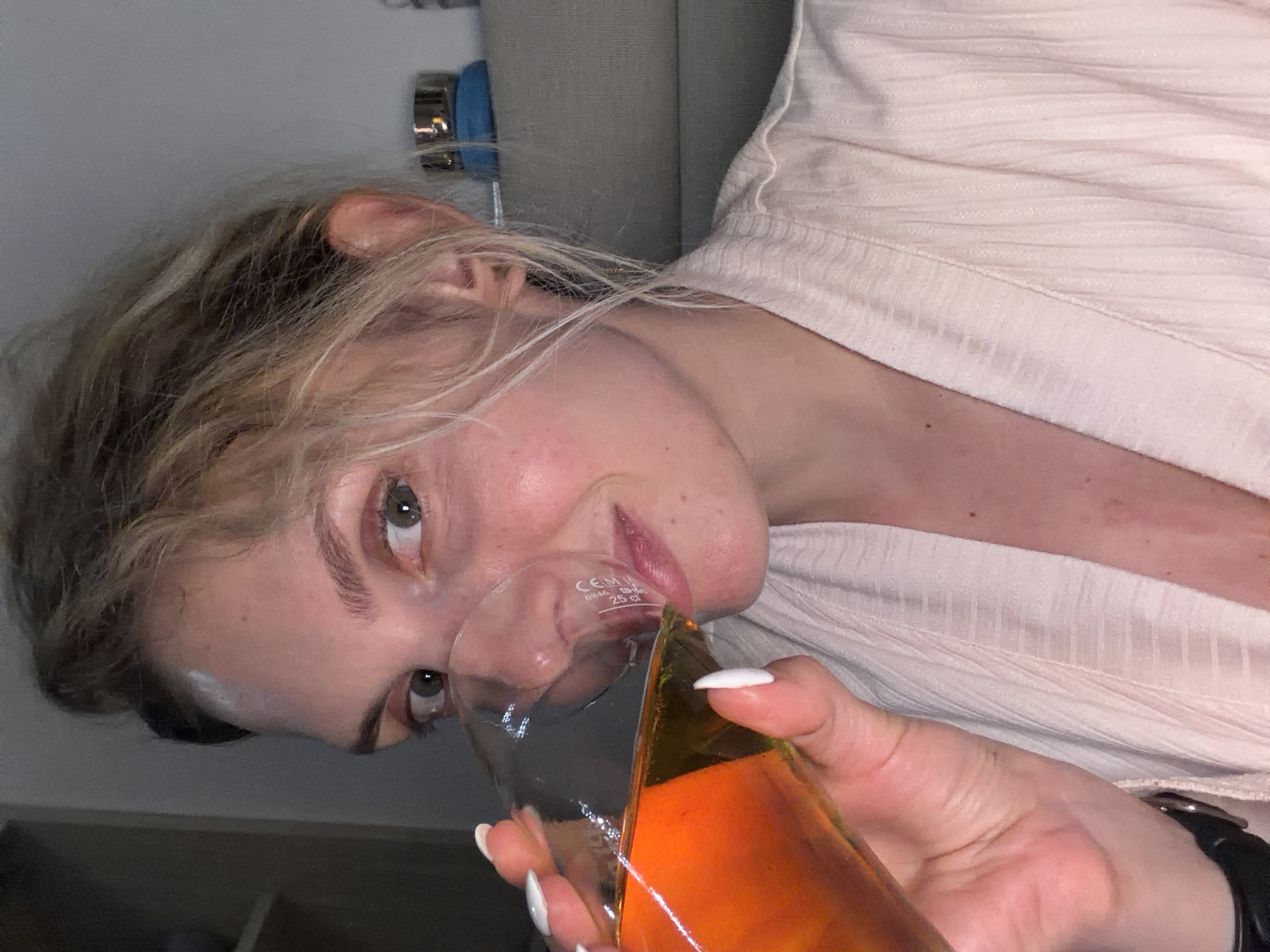 A woman with blond hair and fair skin is seen in a close-up shot, holding a glass containing an orange liquid to her lips. She is wearing a light-colored shirt. The glass appears to be a measuring beaker with markings, suggesting it might be laboratory glassware. The environment is undefined; the background consists of a partial view of what seems to be a wall and furniture. There is no visual indication of Budva, Montenegro, or any other specific location.