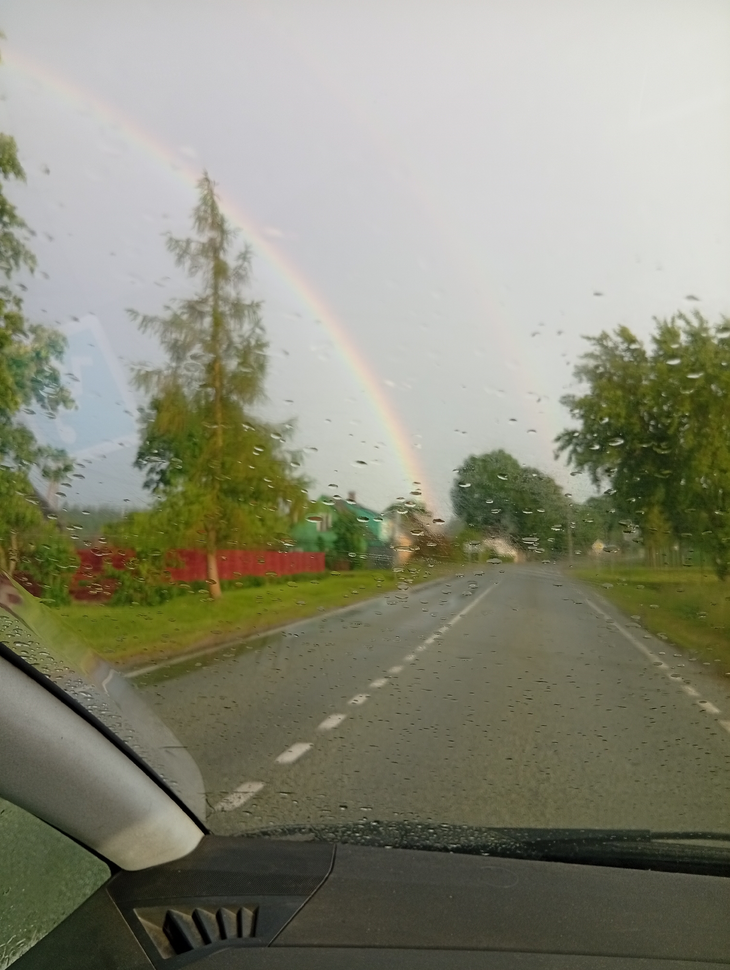 The image, captured from inside a vehicle through a rain-streaked windshield, depicts a rural road scene in Varputėnai, Lithuania. A double rainbow arcs across a pale, overcast sky, with the sky appearing bright and washed out. The road, wet from recent rain, stretches into the distance, marked by white dashed lines. On the left side of the road, a vibrant red fence runs along a grassy verge, bordering a cluster of green trees and a small, green-roofed building. A tall, slender conifer stands prominently near the road. On the right, a dense line of mature green trees flanks the road. The vehicle's dashboard is visible in the foreground, suggesting the image was taken while driving. No people are visible in the scene. The overall impression is one of a post-rain, bucolic landscape.
