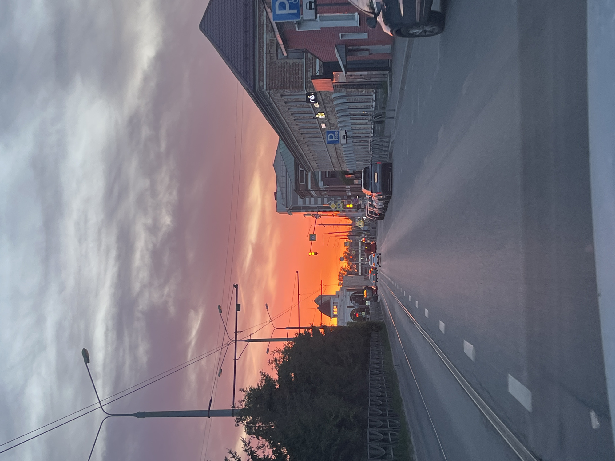 A low-angle view depicts a street in Kazan, Russia at sunset. The sky is a mix of purple, pink, and orange. Buildings line one side of the street, featuring parking signs. Cars are driving on the right side of the street. There are streetlights and utility poles along the street. The road markings include solid white lines. A section of the street has a metal fence. The street is well-lit by the sunset.