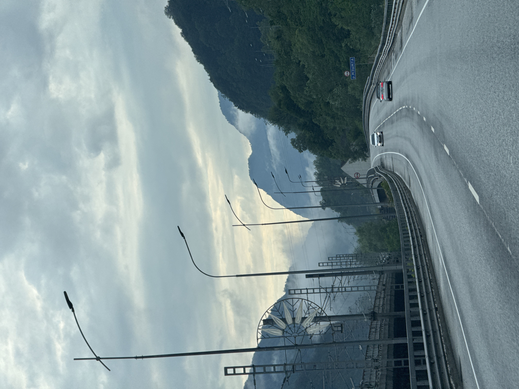 The image shows a highway in a mountainous, cloudy environment. There are several cars on the road, including a white sedan and a silver sedan. The road is flanked by a metal guardrail and on the left side, there is a train track with overhead electrical wires and a decorative, circular structure that resembles a snowflake or sun. Streetlights are also visible along the road. The background is dominated by lush green, tree-covered mountains, with mist or clouds obscuring the higher peaks. A blue road sign with Cyrillic text, "р. МЗЫМТА" (R. Mzymta), indicating a river, is visible on the right side of the road. This location is identified as being in Chvizhepse, Russia, known for its mountainous terrain and proximity to the Mzymta River. The overall atmosphere is overcast and somewhat hazy.