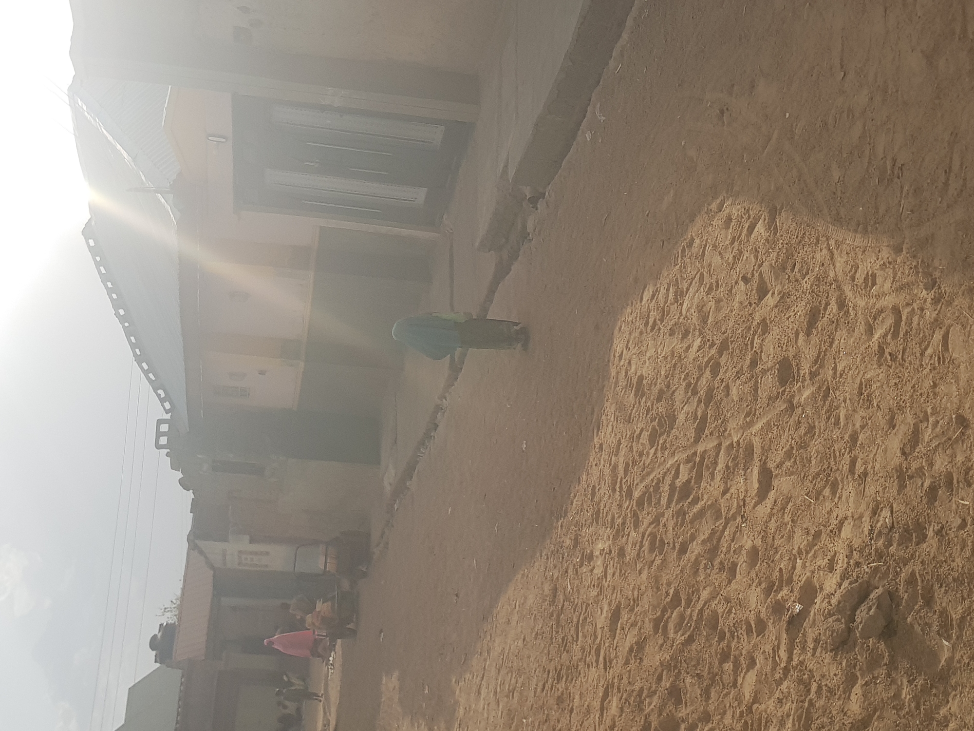A street scene in Dutsinma B, Nigeria. A person in a green garment walks along a dirt road. The road is textured with footprints and various markings. To the left, a building with a corrugated metal roof and a grey facade is present. In the background, there are hints of other buildings and people. The sky is obscured by a bright haze.