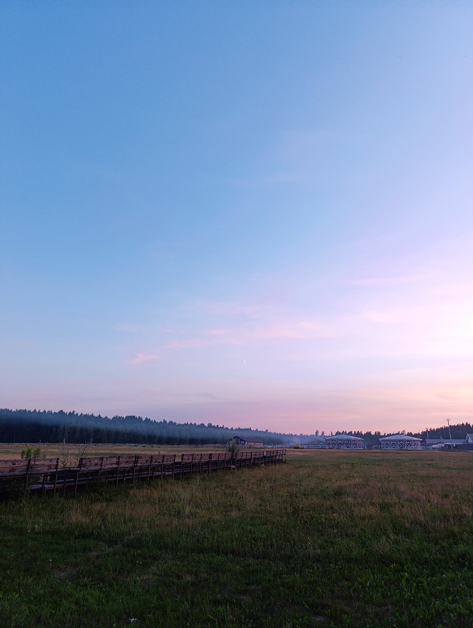 An outdoor, eye-level, wide shot captures a rural scene at dusk. The sky transitions from a clear, light blue at the top to pale pink and then white near the horizon, with a sliver of a moon visible. In the middle ground, a long, wooden fence or railing runs horizontally across the frame, separating a grassy field from a line of trees in the background. Beyond the trees, several white buildings with dark roofs are visible, suggesting a farm or rural settlement. The foreground is a field of tall, green grass.