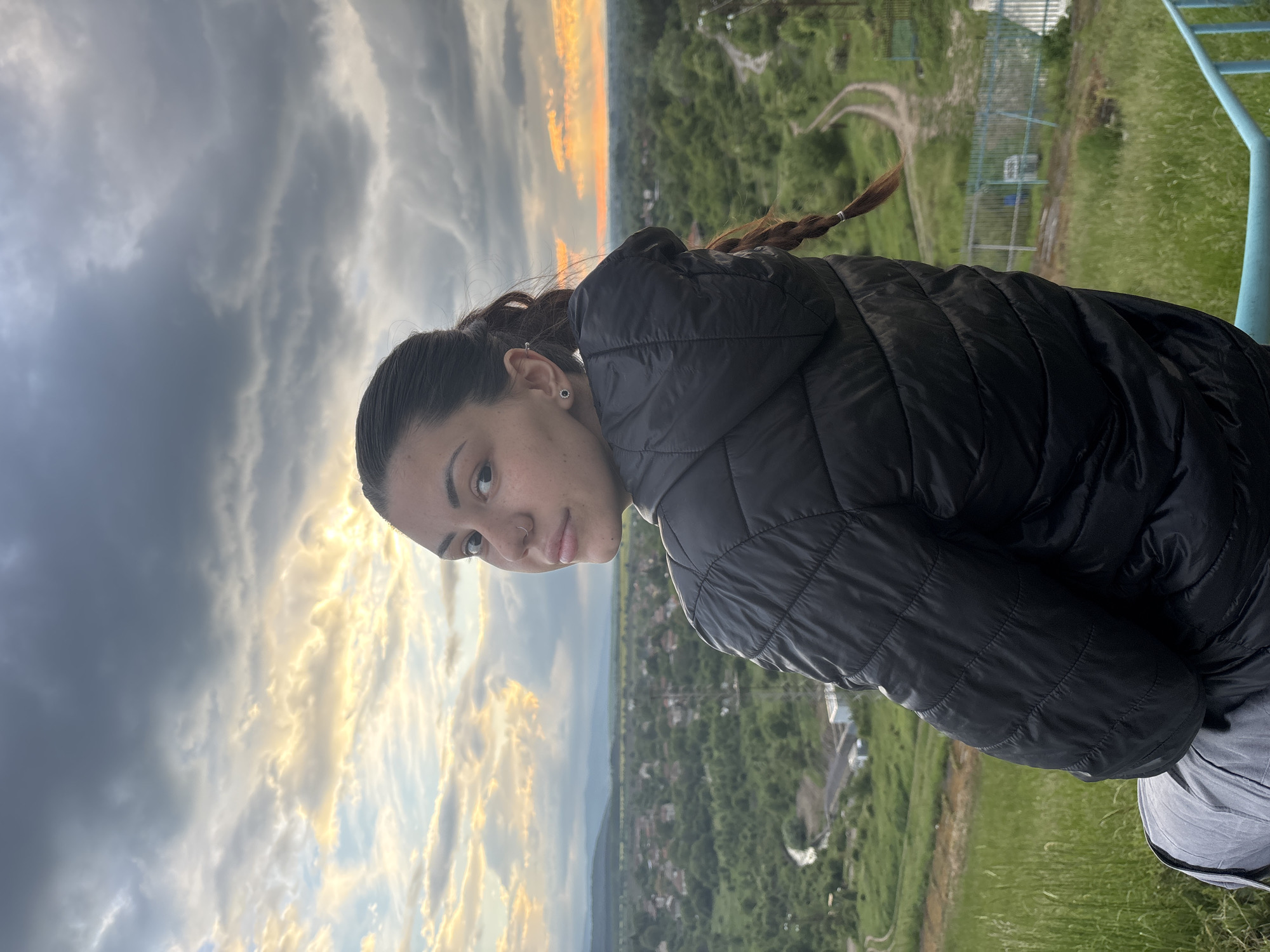 A young woman with brown hair pulled back in a ponytail and a black puffer jacket poses outdoors. She is looking at the camera with a slight smile. Her left ear has an earring and her left nostril has a nose ring. In the background, there is a cityscape with a green landscape and a sky with dramatic clouds at sunset. The scene takes place in Ognyanovo, Bulgaria, as seen from an elevated position overlooking the city. A light blue metal rail is visible in the extreme upper right corner of the image.