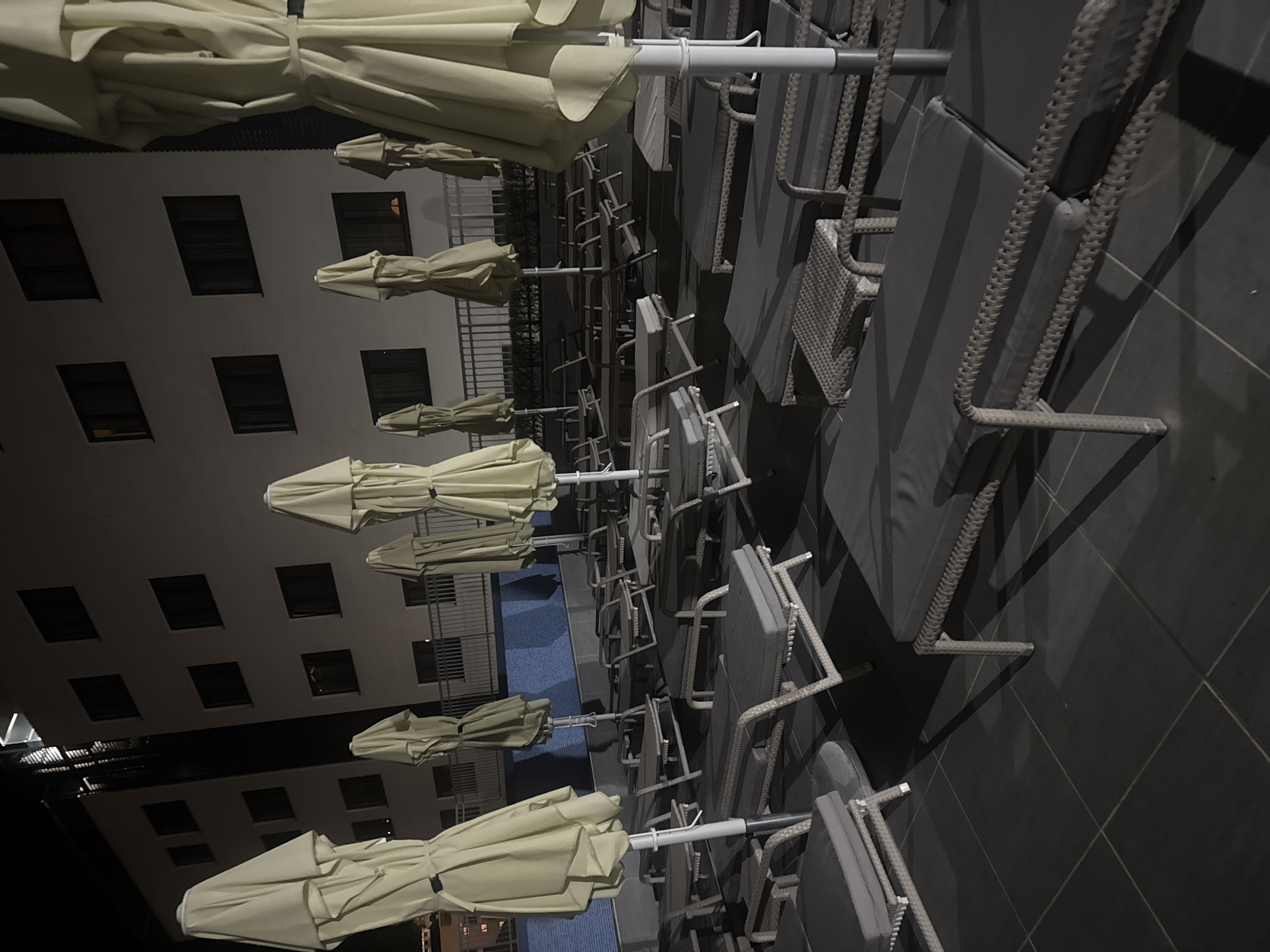 The image shows an outdoor space at night in Unknown, Unknown. Rows of closed, light-beige patio umbrellas and lounge chairs are arranged along the edge of a blue-tiled pool. A multi-story building with evenly spaced, dark windows forms the backdrop. The pool area is lit, but the scene has a somber tone due to the time of day.