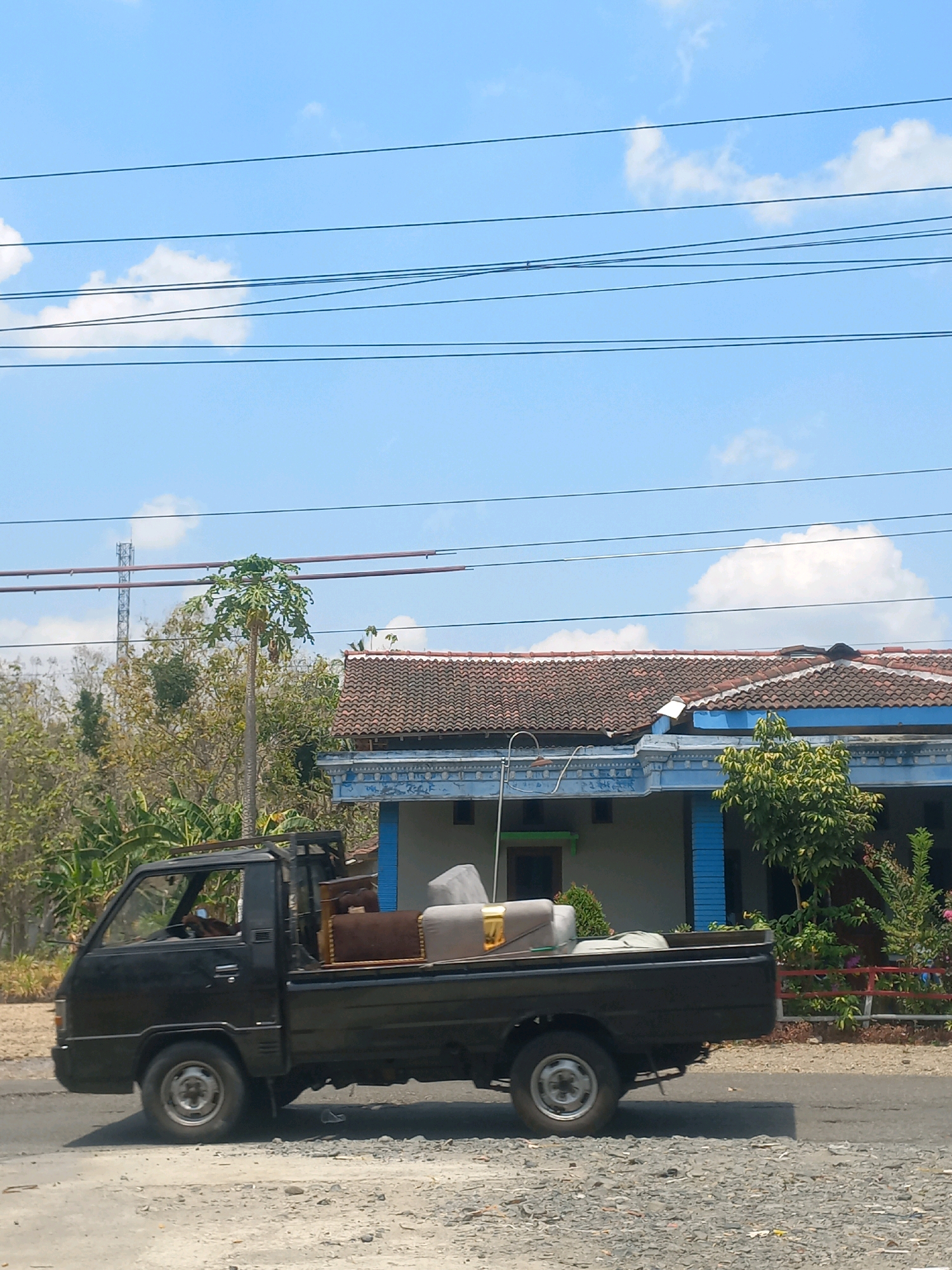 A black pickup truck is traveling on a paved road, carrying furniture in its open bed. The truck has two visible wheels on its side. In the background, a residential building with a tiled roof and blue trim is visible, with trees and foliage surrounding it. A cell tower can be seen in the distance. The sky is blue with scattered white clouds, and power lines are strung across the top of the image. The scene appears to be in Baturetno, Indonesia, given the architectural style and vegetation.