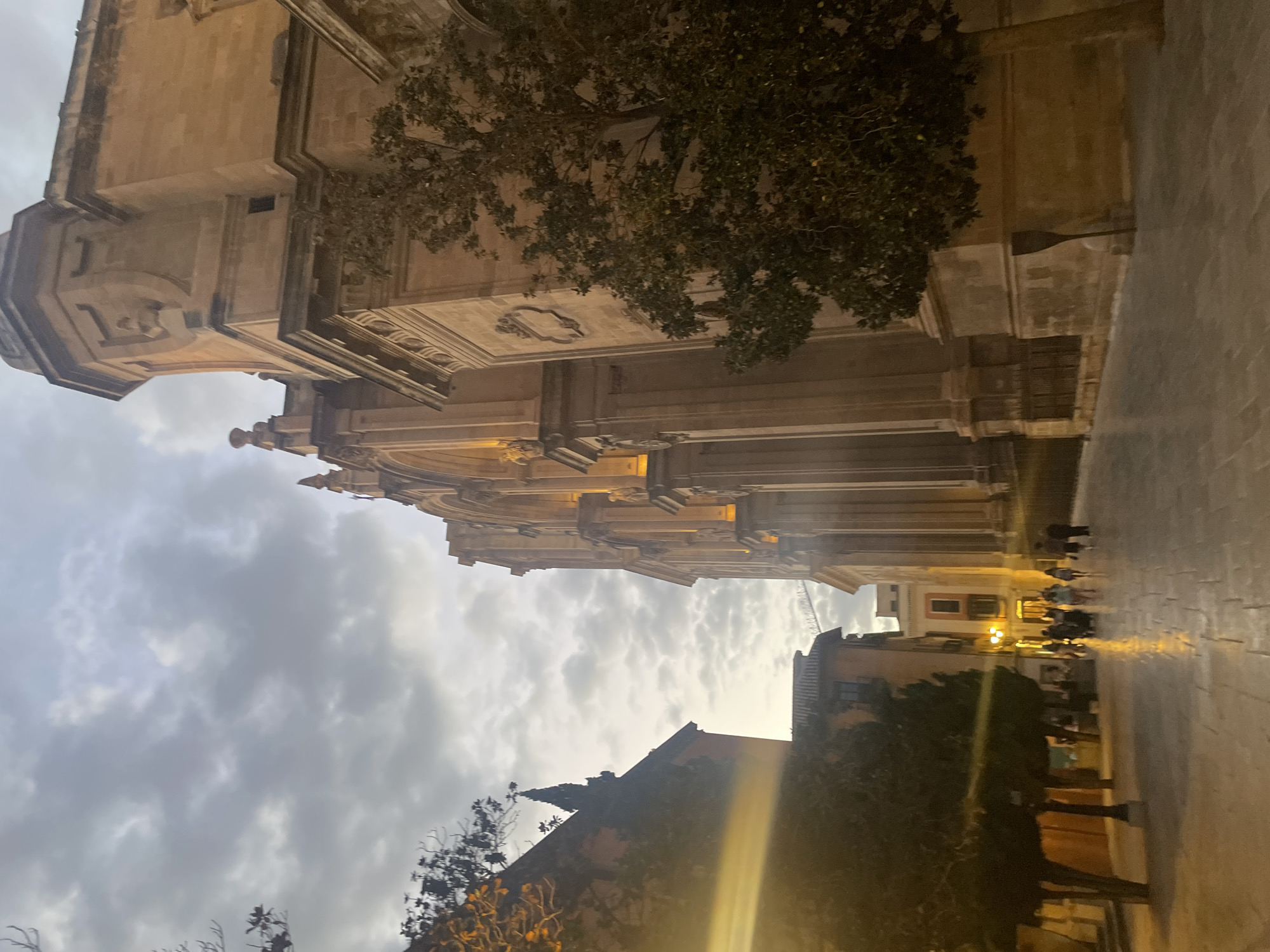 The image depicts the Granada Cathedral in Granada, Spain, at dusk. The grand facade of the cathedral, constructed from tan stone, dominates the right side of the frame, characterized by its imposing columns, arches, and decorative elements. A tree with dense green foliage stands in front of the cathedral, partially obscuring its lower section. The sky above is filled with dramatic, grey clouds, suggesting overcast weather or the transition from day to night.
To the left of the cathedral, another building, appearing older and with a reddish hue, is visible, its architectural style contrasting with the cathedral's grandeur. Trees with sparse branches are planted along the walkway. The ground is paved with cobblestones, which reflect the ambient light, creating a glistening effect, especially further down the path.
A group of people is walking away from the viewer, their figures silhouetted against the illuminated buildings in the background. The lighting suggests streetlights and interior lights from nearby establishments are on, casting a warm glow on the scene. The overall atmosphere is one of serene, historic urban twilight.