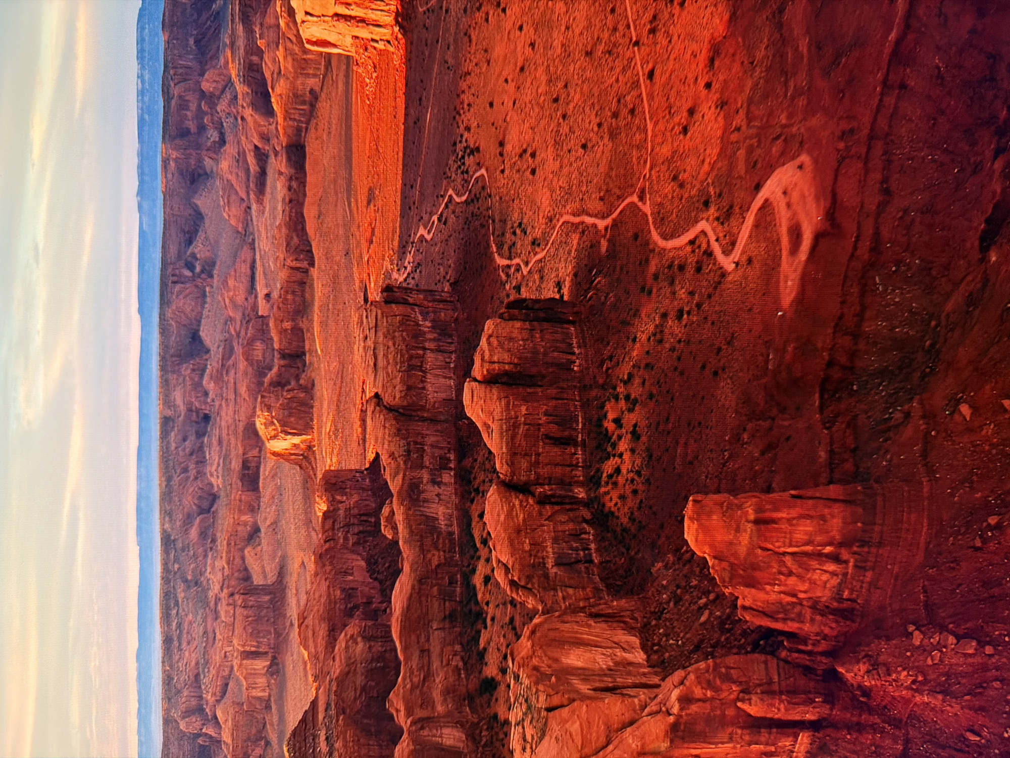 The image depicts a wide, panoramic view of a desert landscape, characterized by towering red rock formations, vast expanses of arid terrain, and a winding dirt path. The scene is illuminated by the warm, golden light of either sunrise or sunset, casting deep shadows and highlighting the vibrant red and orange hues of the rocks and earth. In the distance, a hazy blue mountain range is visible against a pale sky with wispy clouds. The formations are layered and sculpted, suggesting geological erosion over a long period. The terrain is sparsely vegetated with low-lying shrubs and scattered rocks. A narrow, winding path traverses the landscape, indicating a human-made trail through the wilderness. The perspective is aerial or from a high vantage point, allowing for a comprehensive view of the vast and rugged environment. The scene evokes a sense of remoteness and natural grandeur. The location is not in Kyiv, Ukraine.