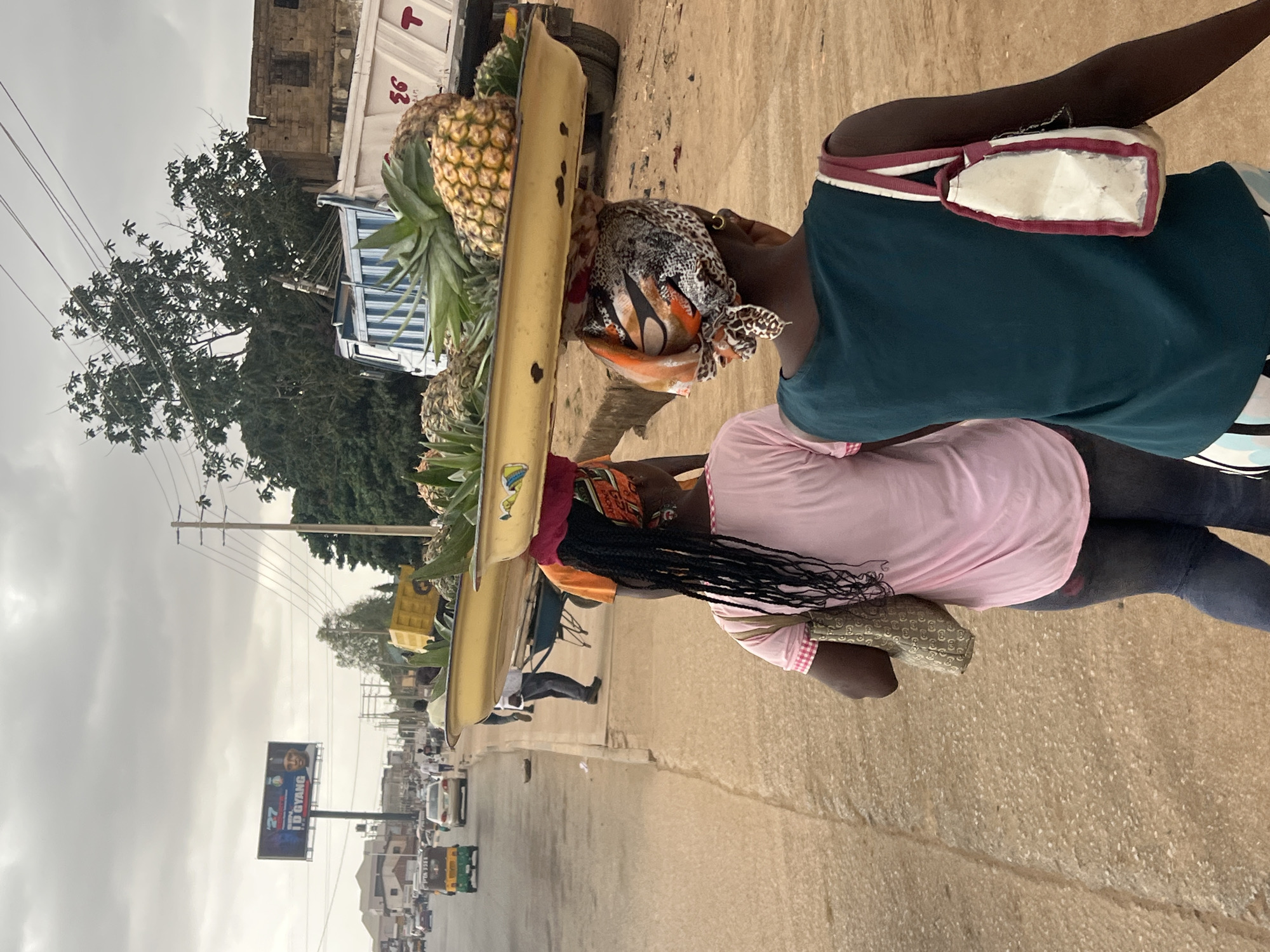 The image, rotated 90 degrees clockwise, captures a street scene in Gura Top, Nigeria, featuring two women in the foreground on a dirt roadside next to an asphalt road. The woman closer to the viewer wears a teal sleeveless top and dark trousers, with a white and maroon strap bag slung over her left shoulder. She carries a long, light yellow tray on her head, laden with multiple ripe pineapples. The second woman, slightly behind and to her left, wears a pink top and dark trousers, with a patterned purse on her right arm. Her long, black braids extend down her back.
The street, which appears busy, stretches into the background, populated by various vehicles including cars, auto-rickshaws (keke napep), and trucks. One white and blue truck is partially visible behind the women, and another yellow/tan truck is further down the road. Buildings line both sides of the street. On the left side of the road, a large billboard is prominent, displaying "27 TD GYANG" with an image of a man. Utility poles with power lines run along the street. The sky is overcast, suggesting a cloudy day, and the overall lighting is diffuse. The scene depicts daily life and commerce in an urban or semi-urban environment. Visible text includes "27 TD GYANG" on the billboard and "93" on the side of a white truck, along with a red "T" logo. The patterned bag carried by the woman in pink appears to have the letters "G UCCI".