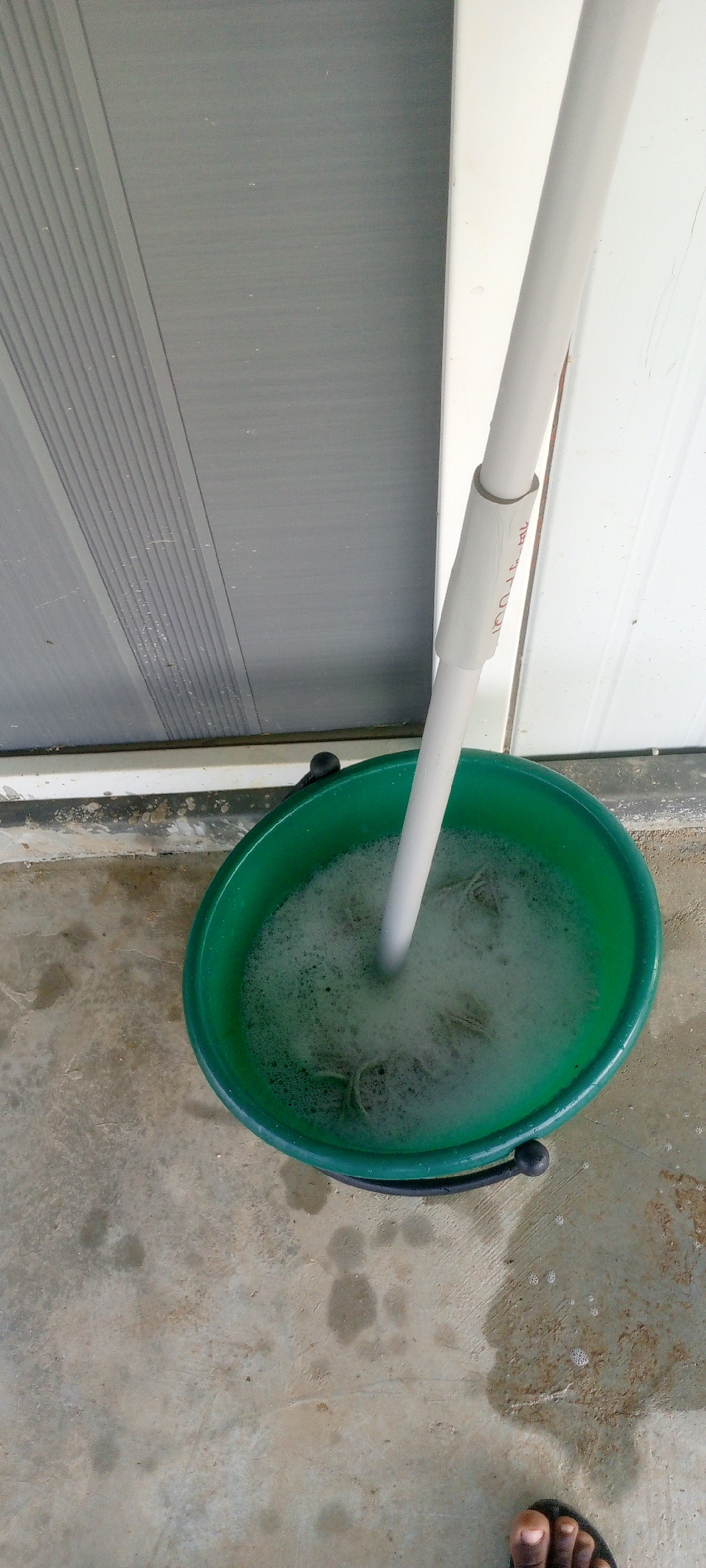 A close-up, overhead view shows a green plastic bucket filled with soapy water and a mop. The bucket is on a concrete surface in front of a grey, ribbed door and a white vertical structure. A white pole, likely the handle of the mop, extends from the bucket and upwards out of the frame. Some of the mop head is submerged in the soapy water, with visible suds and strands of the mop. The concrete surface appears wet in places and shows some dirt. A person's bare foot wearing a black sandal is visible in the bottom right corner of the image. The environment suggests an outdoor or semi-outdoor location, possibly a patio or entryway. The scene is located in Indo, Nigeria.