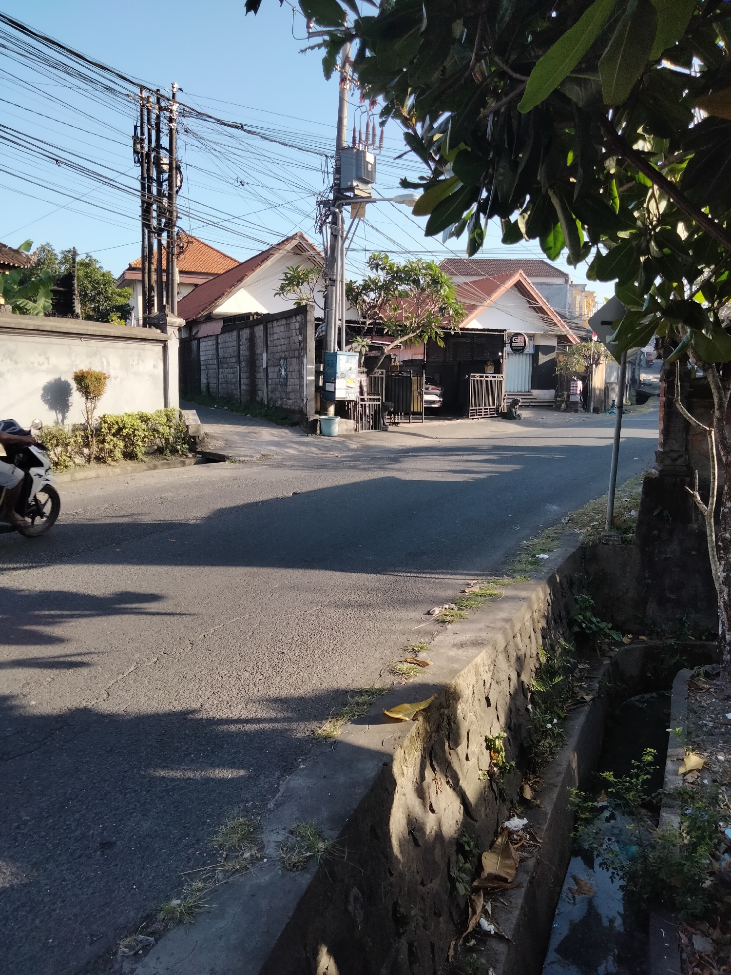The image depicts a street scene in Denpasar, Indonesia, bathed in the warm, long shadows of late afternoon or early morning sunlight. The road surface is asphalt, with a visible concrete drainage ditch running along the right side of the frame, filled with murky water and debris. A low, stone-walled embankment borders the ditch.
On the left side of the road, a light-colored motorcycle with a rider, partially visible, is moving away from the camera, casting a long shadow on the asphalt. Adjacent to the road on the left is a light-colored concrete wall, behind which are some neatly trimmed bushes.
Further down the street, buildings with terracotta tile roofs are visible. One building features a dark-colored facade with a metal gate, and another displays a white facade with the letter "G" in a black circle. Utility poles with a dense network of electrical wires crisscross the sky above the buildings and the street. Trees with broad green leaves are present on both sides of the street, with some branches obscuring the view of the sky. The overall environment suggests a residential or mixed-use urban area with typical Indonesian infrastructure.