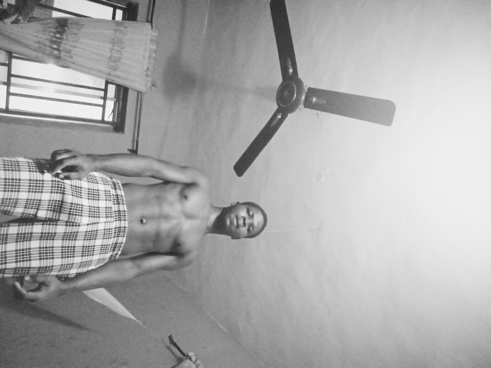A young Black male, shirtless and wearing plaid shorts, stands in a dimly lit room. He is facing the camera with his head tilted slightly upwards. His abdominal muscles are visible. Above him, a ceiling fan with three blades is mounted on the ceiling. To his left, a window with a patterned curtain is visible. The room appears to be a bedroom or living space. The photograph is taken from a low angle. The image is in black and white and has a grainy texture.