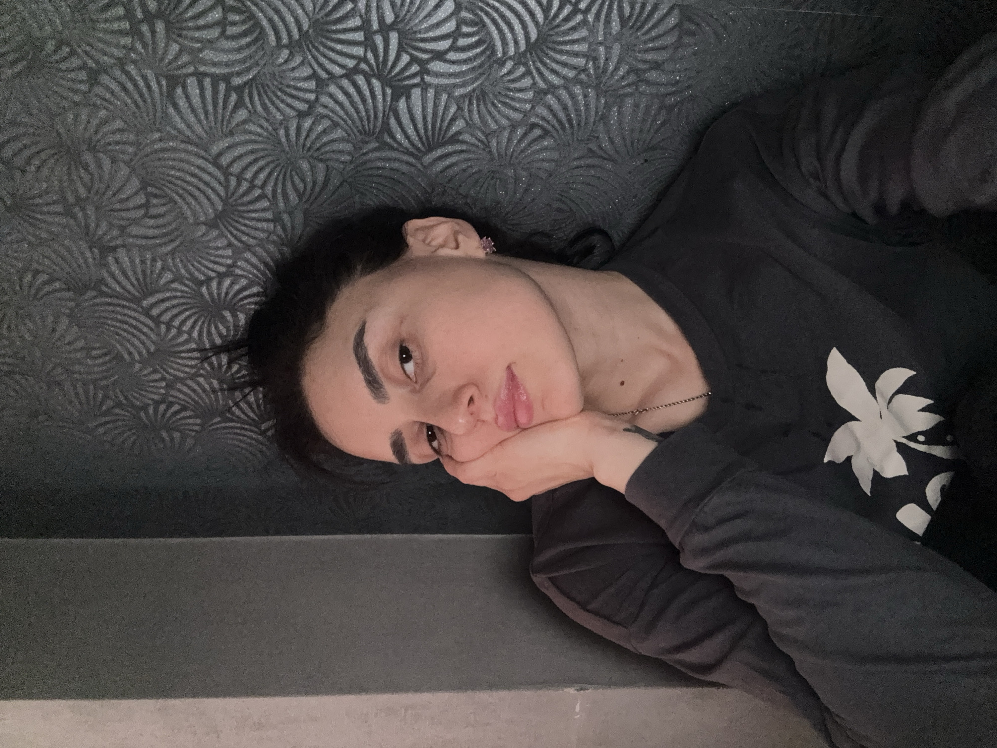 A person with dark hair is lying down on a grey surface. Their head rests on their hand. The person is wearing a dark grey long-sleeved shirt with a white floral design on the front. Behind the person is a wall with a patterned design that resembles repeating fans or shells. The exact location in Tbilisi, Georgia is not discernible from the image.
