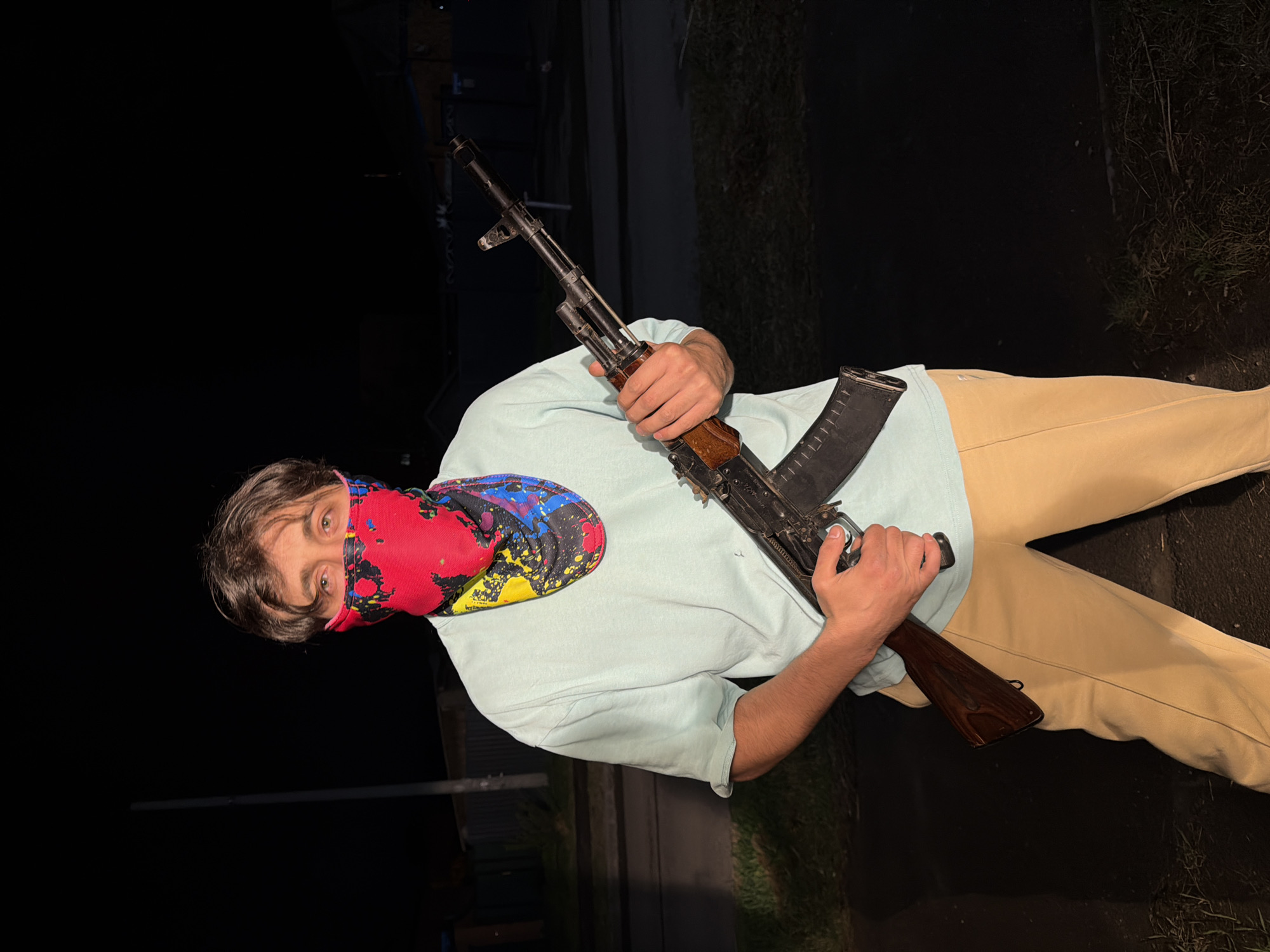 A person with brown hair and eyes, wearing a red, blue, and yellow patterned face covering, is holding a rifle. They are wearing a light blue t-shirt and khaki pants. The background appears to be dark, possibly at night, with some vegetation and structures visible. The person is standing on a paved surface. There are no additional details indicating the location in Beryozovka, Russia.