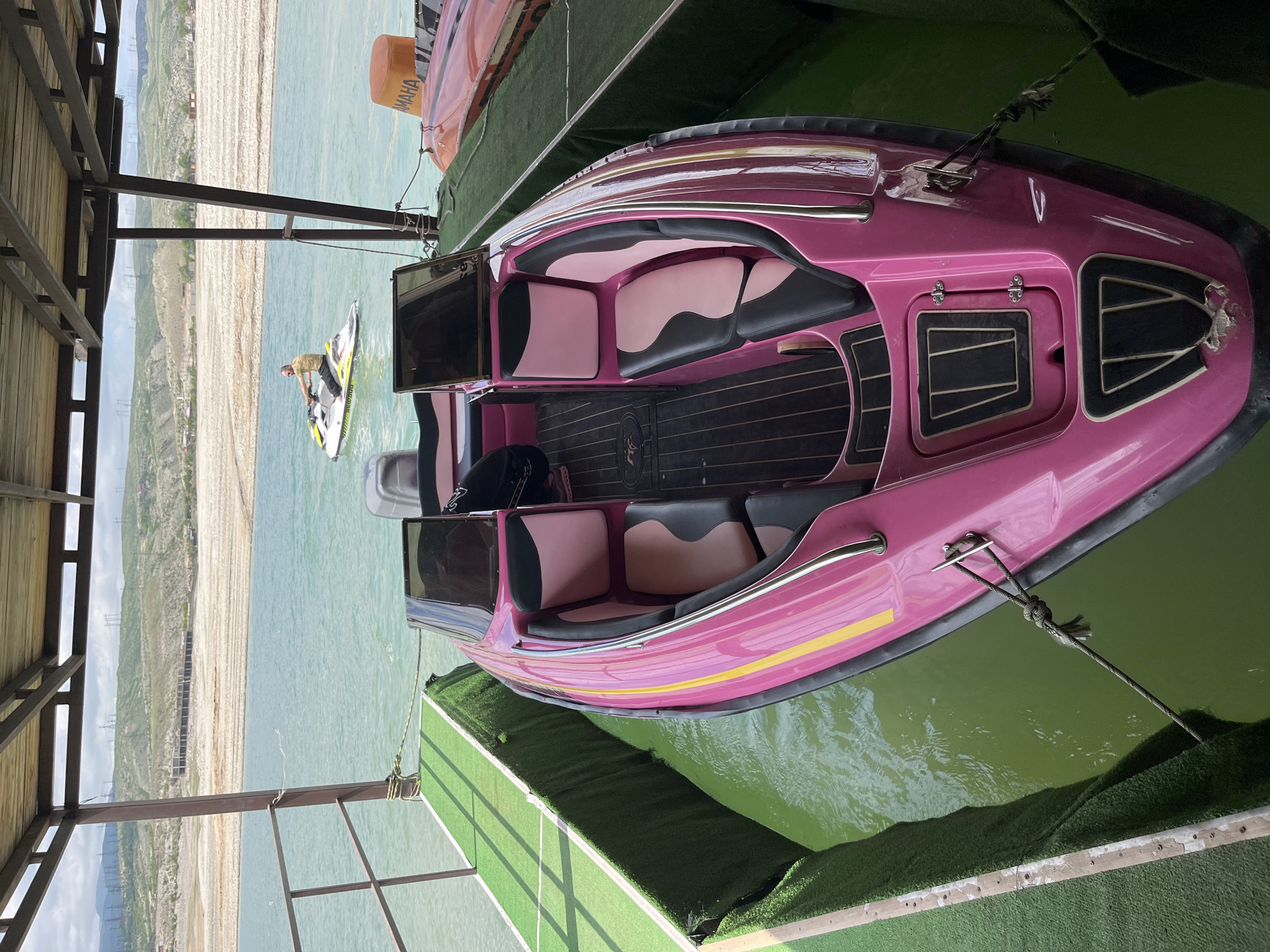 The image depicts a pink speedboat docked on a floating platform covered in artificial turf, with a view of the Chirkey reservoir, located near the city of Chirkey, Russia. The speedboat features a black interior with pink and black seats. A personal watercraft with a person aboard is visible in the distance on the reservoir. A second watercraft rests on the platform behind the speedboat. A covered structure, likely a dock, is visible on the left side of the image. The reservoir shore appears arid with signs of erosion.