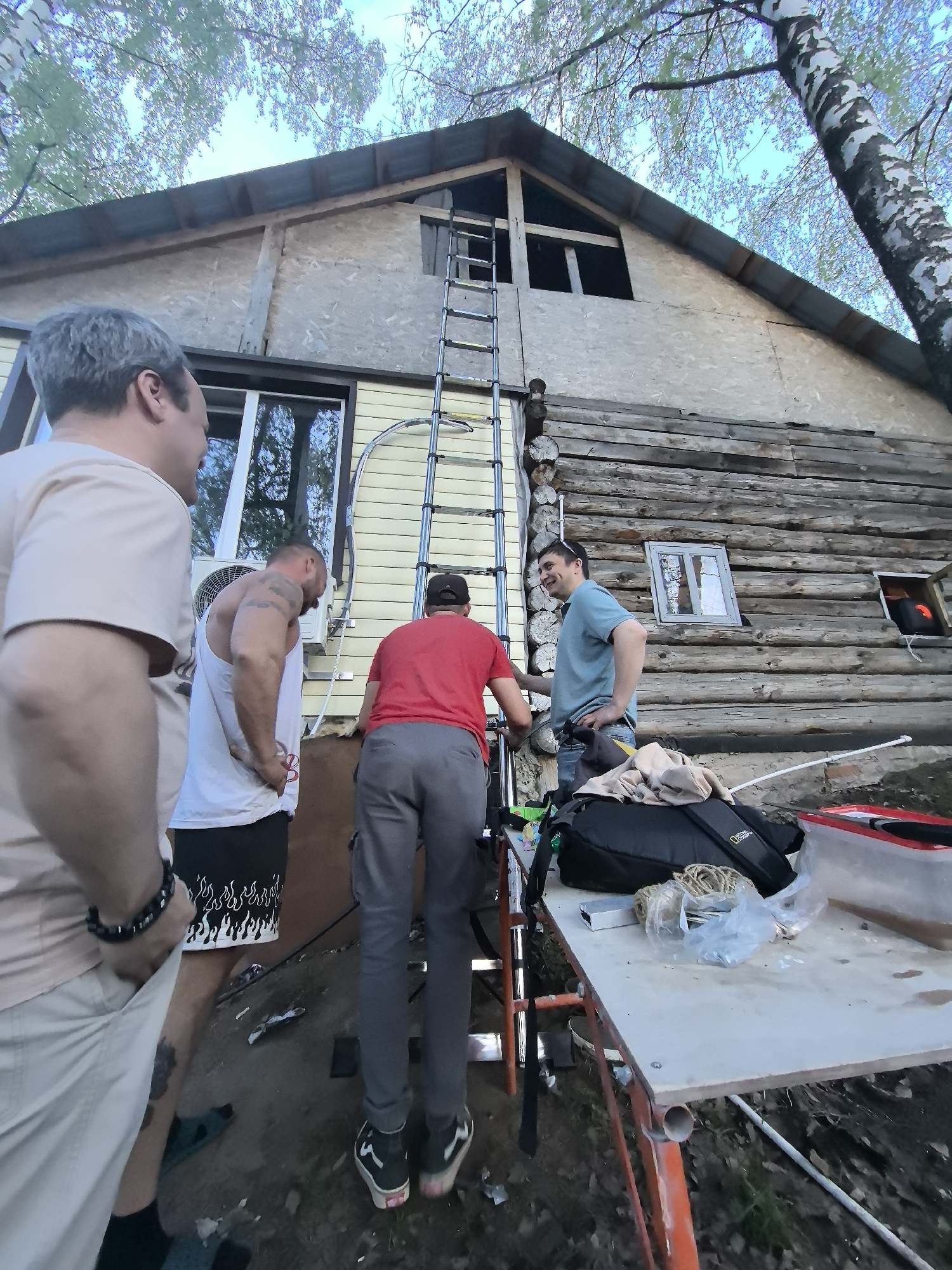 Four adult males are gathered outdoors in Noviy, Russia, in front of a two-story building. The building has a mixed facade: the lower portion is constructed from stacked logs, while the upper section is covered in light-yellow paneling. A tall, silver ladder leans against the building, reaching towards a triangular window near the roof. A table, assembled with orange scaffolding, is set up to the right of the ladder. Items such as a backpack, various bags, and a red container are placed on the table. The men are dressed in casual attire. One man is wearing a light-colored shirt, black shorts with a white flame pattern, and sandals. Another is clad in a red shirt and gray pants, standing near the base of the ladder. A third man, wearing a light-blue shirt and jeans, is standing to the right, facing the other men. The fourth man is on the left. A birch tree stands near the building. An air conditioning unit is mounted on the yellow-paneled wall.