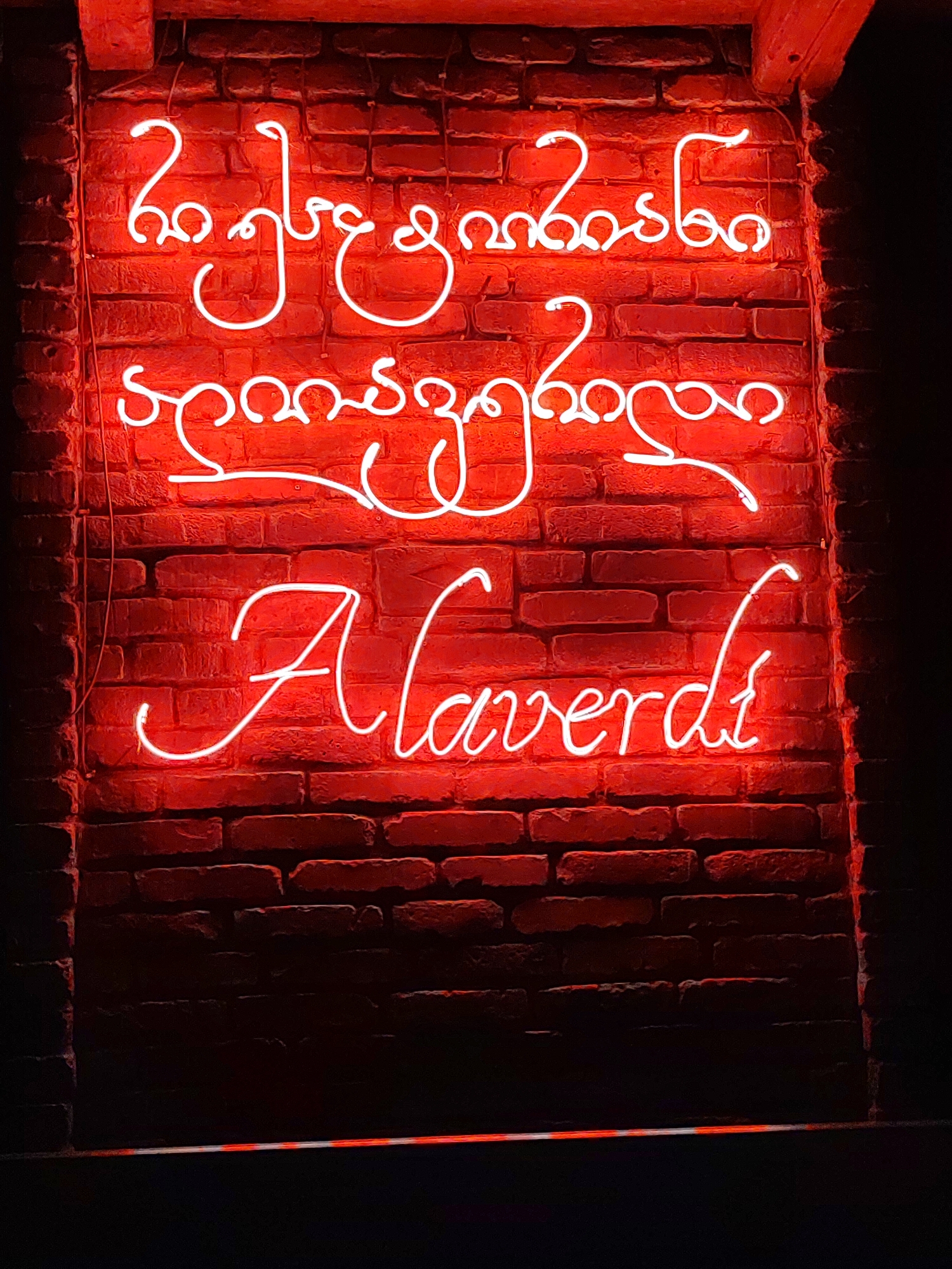A red neon sign with white lettering is mounted on a brick wall. The sign has two lines of text in Georgian script followed by one line in Latin script. The Georgian script translates to "Georgian wine," and the Latin script spells out "Alaverdi," which is a type of Georgian wine. The brick wall is dark red and appears to be made of weathered bricks. The lighting is dim, with the neon sign being the primary light source. The bottom of the image shows a dark area, possibly a floor or a table, with a faint red glow from the neon sign.