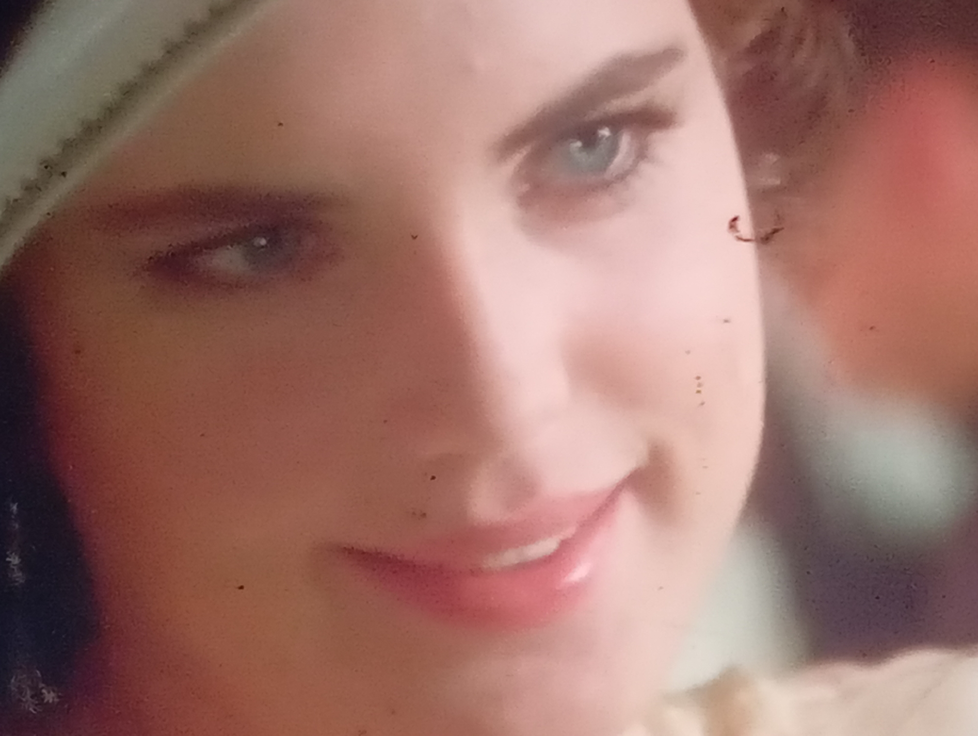 The image is a close-up portrait of a young woman's face. Her right eye is visible, a light blue iris with a pupil. Her mouth is slightly open, revealing teeth, and her lips are painted a glossy pink. A portion of her forehead and cheek are also visible. The background is out of focus and appears to be a soft, warm color. There are several small dark specks on the image, likely dust or imperfections in the photograph. The overall impression is an intimate and slightly stylized portrait. The image is not a scene from Sofiivska Borshchahivka, Ukraine; it is a close-up of a face.