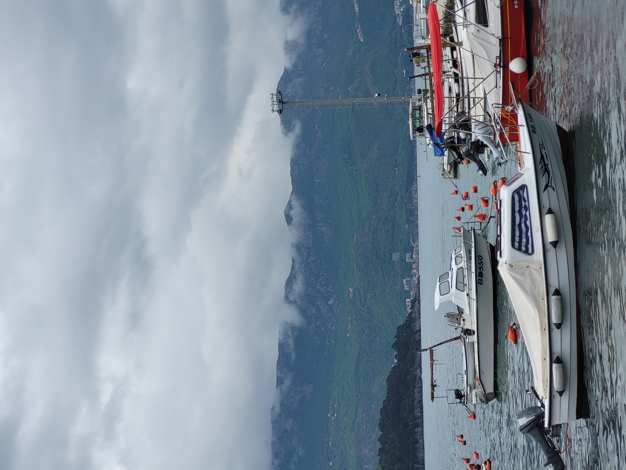 This image captures a tranquil harbor scene in what is identified as Budva, Montenegro. The view looks across a body of water towards a dramatic mountainous coastline partially shrouded in clouds.
In the foreground and midground, several boats are moored. Closest to the viewer is a white boat with dark blue striped cushioning visible on its canopy. It features a distinctive black fish graphic on its side and white fenders along its hull. Behind it, another white boat is prominent, marked with "BD 550" and equipped with a "HONDA" outboard motor. A "SUZUKI" outboard motor is also visible on this vessel. Further back, part of a larger red and white boat can be seen, with a vibrant red canvas cover. Numerous orange and white buoys or fenders float in the water around the boats.
The background is dominated by a majestic mountain range, lush with green vegetation. The peaks and upper slopes are partially obscured by low-hanging gray clouds or mist, creating a moody atmosphere. Scattered buildings are visible on the lower mountain slopes, indicating coastal settlements. A tall, slender metal tower structure stands on the shoreline or lower mountain, possibly for communication or navigation.
The sky is overcast with dense gray clouds, suggesting a cloudy day. No people are visible, contributing to the quiet and serene ambiance of the scene. The boats appear to be at rest, with no discernible activity taking place. The overall impression is one of a peaceful coastal harbor set against a rugged, natural backdrop.