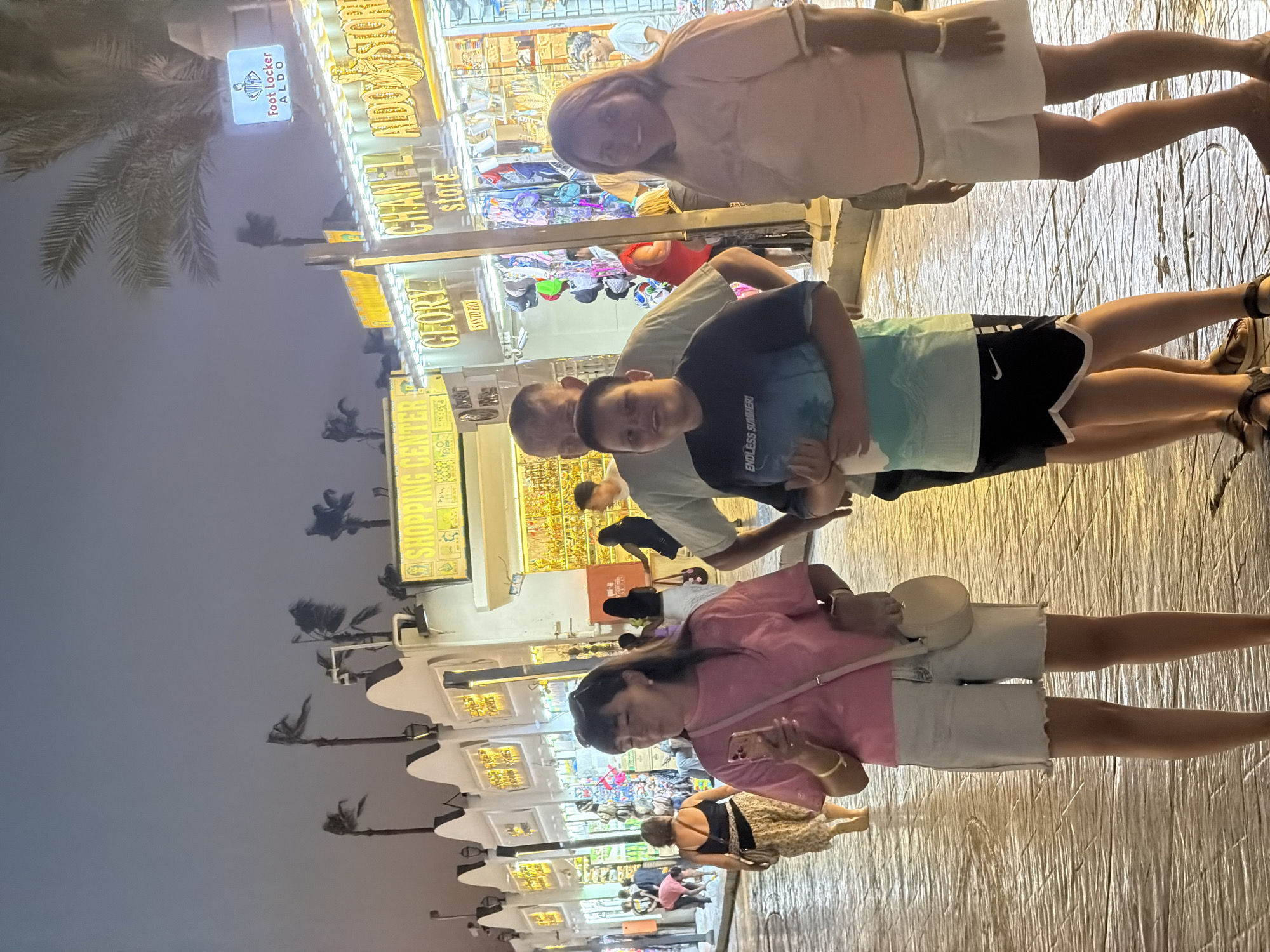 The image depicts a street scene at night in Sharm El Sheikh, Egypt, likely in a commercial or tourist area. The foreground features a family group of four, with two adults and two children. A woman in a pink t-shirt and white shorts holds a phone in her left hand and wears a white crossbody bag. To her right, a young boy in a dark t-shirt with a faded graphic print and black shorts stands next to a man in a light-colored t-shirt. A young girl in a light pink t-shirt and white skirt stands on the far right.
In the background, a row of shops with illuminated signs lines the street. Visible signs include "SHOPPING CENTER," "GLORY," and "CHANEL store." One sign also indicates "ALDO STORE." The architecture appears to be of Mediterranean or Middle Eastern influence, with white facades and decorative elements. Palm trees, some with fronds bent by wind, are visible against the dimly lit sky. The ground is paved with stone or brickwork that appears wet, possibly from recent rain or cleaning. Several other individuals are visible walking in the background, suggesting a public thoroughfare. The overall atmosphere is lively and commercial, characteristic of a popular tourist destination.