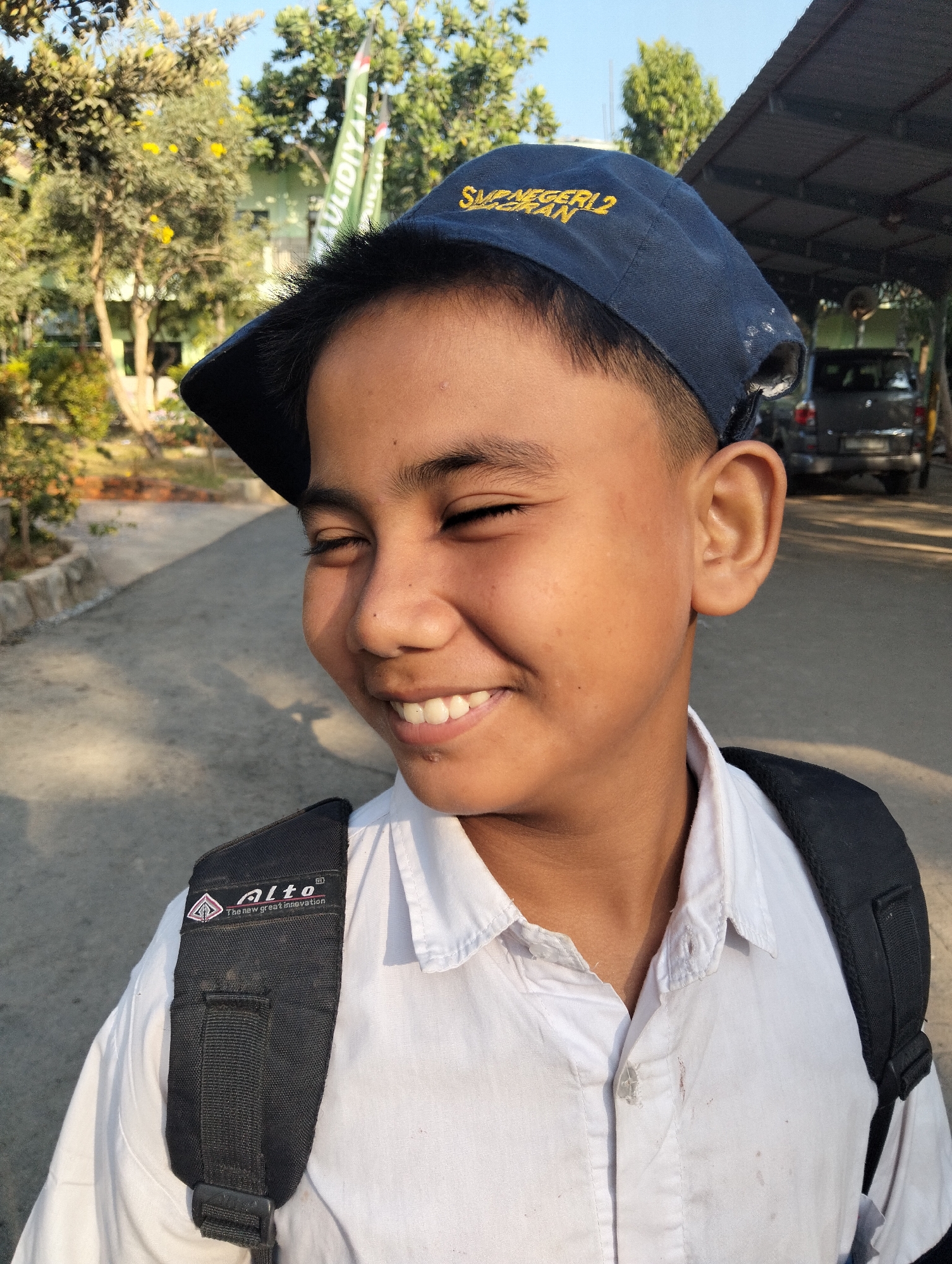 A young male, appearing to be of school age, is captured in a close-up portrait outdoors. He is wearing a blue baseball cap with yellow lettering that reads "SMP NEGERI 2" and "TERAN," indicating he attends a junior high school. He also wears a white collared shirt and a black backpack with the "Alto" logo visible on one of the straps. The boy is smiling with his eyes closed, suggesting a moment of happiness or amusement.
The background reveals an outdoor setting with trees and vegetation. A paved path is visible behind the boy, leading towards a covered structure, possibly a carport or pavilion, under which a dark-colored vehicle is parked. Sunlight casts shadows on the ground, indicating it is daytime. The presence of school-related insignia on his cap and the casual outdoor setting suggest this photograph might have been taken near a school in the city of Paciran, Indonesia, possibly after school hours or during a school event.