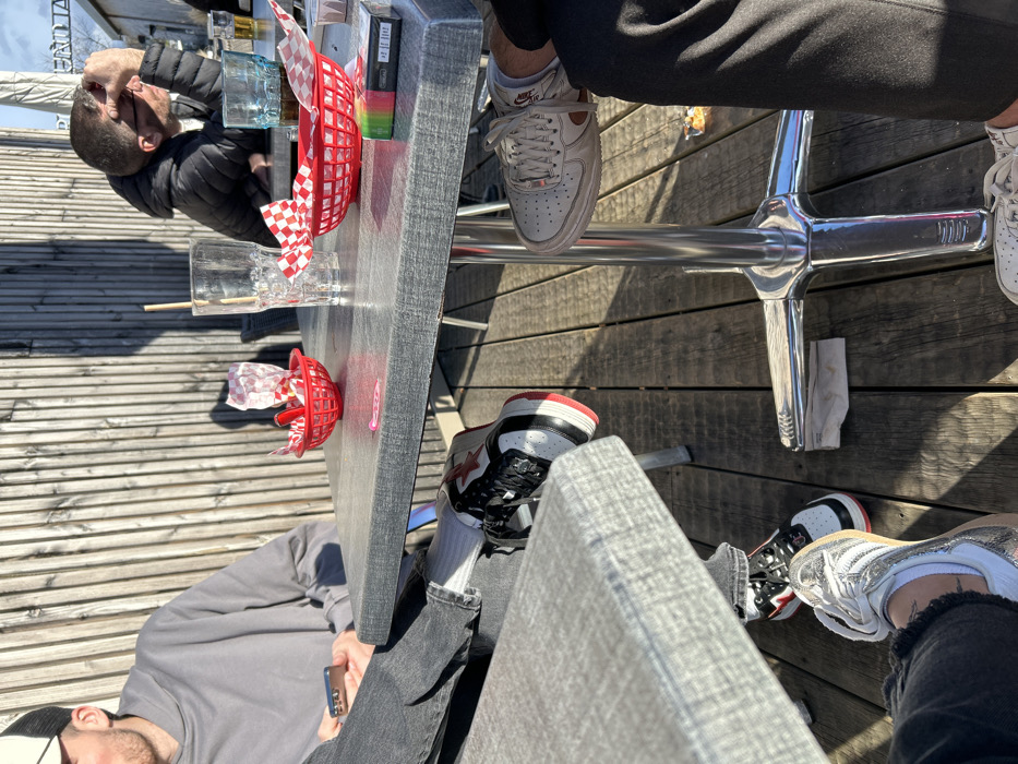 The image captures a casual outdoor gathering, likely on a sunny day, at a table with several individuals present. The perspective is from slightly above the table, looking down at the people's legs and feet, and the items on the table. The ground consists of wooden deck planks, indicating an outdoor patio or similar setting.
Four individuals are identifiable, primarily by their presence around the table and their footwear.
1. On the far side of the table, a person wearing a black puffer jacket is visible, looking down or away.
2. On the near side of the table, another person, wearing a grey long-sleeved shirt and dark jeans, is seated, holding a smartphone in their hands. This person is also wearing a black and white baseball cap.
3. Under the table, the legs and feet of a person wearing black trousers and white Nike Air Force 1 sneakers are prominent. The Nike logo is visible on the side of the shoe.
4. Also under the table, the feet of another person are visible, wearing black, white, and red high-top sneakers, possibly with a star logo on the side, and white socks. Next to these, another pair of white and silver sneakers are also visible.
The table itself has a grey, textured surface and is supported by a shiny, chrome-like pedestal base. Scattered on the table are several items:
* Two red plastic baskets commonly used for serving food like fries or appetizers. One basket contains a red and white checkered paper liner, while the other holds some crumpled paper.
* A clear drinking glass with ice and a wooden stirrer.
* Another glass, partially visible, appears to contain a bluish liquid.
* A black rectangular packet, possibly cigarettes or a vape pen.
* A small, colorful item, possibly a lighter or another small package.
* A small pink sticky note or sticker.
The scene is brightly lit by natural sunlight, casting strong shadows on the wooden deck planks and under the table, suggesting a clear day. The overall atmosphere appears relaxed and social, with people enjoying food or drinks outdoors. No specific emotions are discernible from the partial views of the individuals. No legible text, other than brand logos on shoes, is visible in the image.