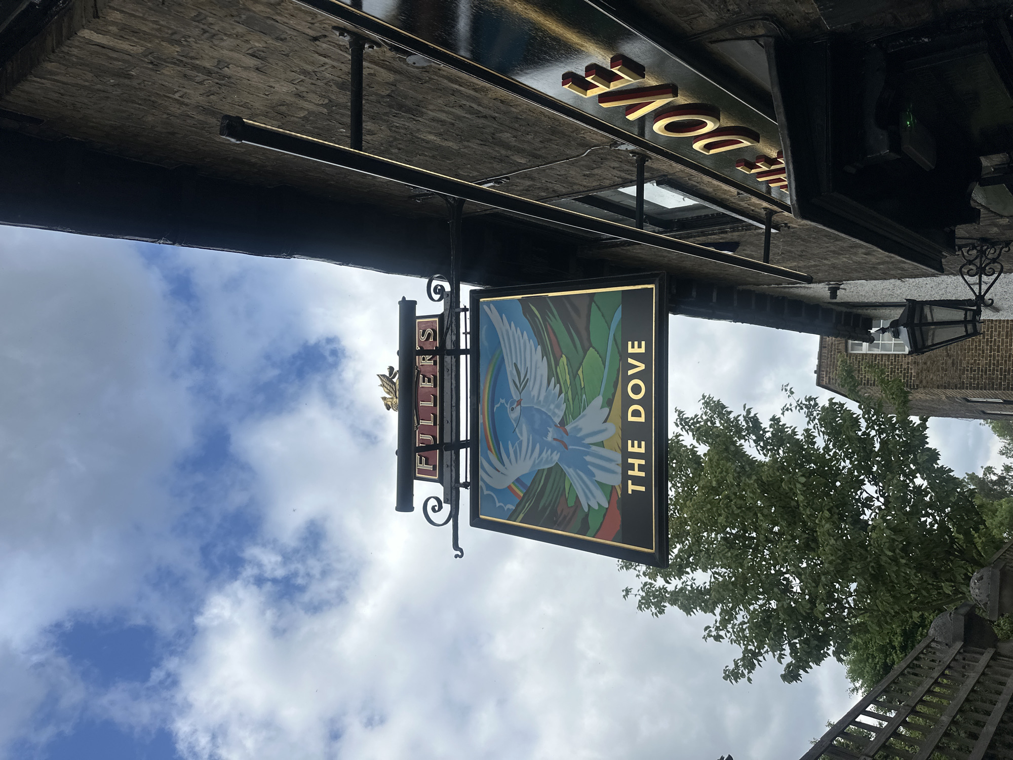 The image shows a pub sign for "The Dove", located in London, United Kingdom. The sign features a white dove illustration against a green and blue background. The word "Dove" is vertically aligned on the sign in gold lettering. The sign also has the word "Fullers" on the left side of the bracket, in gold lettering. The pub is a Fuller's pub. The sky is visible in the background, with white clouds. The image is taken from a low angle, looking up at the sign. The architecture of the pub is visible above the sign, along with a section of a brick wall and a vintage-style lamppost. A tree with green foliage is to the right of the pub sign. A piece of fencing is also visible to the bottom right.