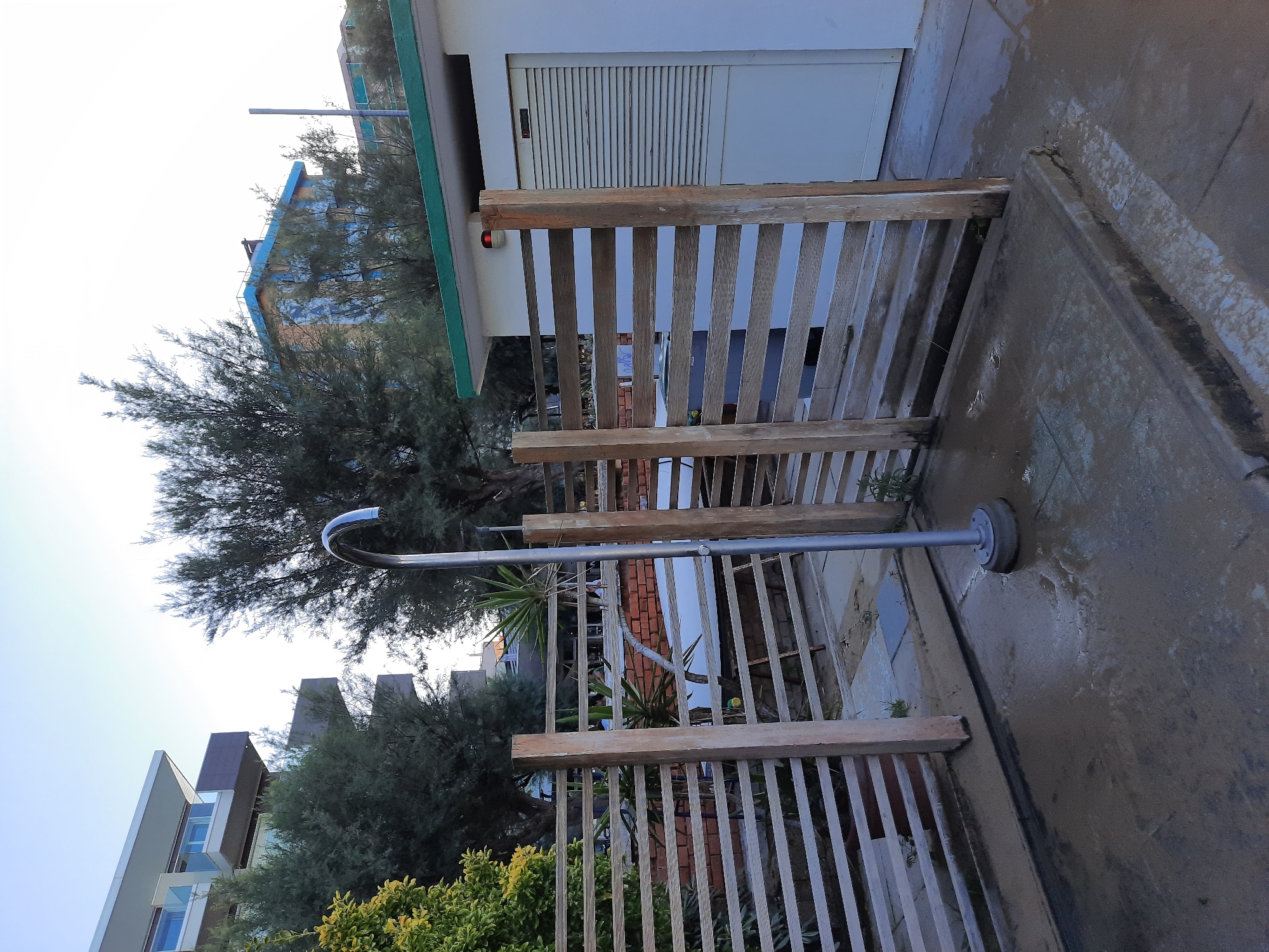 The image shows an outdoor shower stall on a paved area. The stall is constructed with wooden posts and slatted horizontal fencing. A metal showerhead on a curved arm extends from a central pole. The ground around the shower is wet and appears to be sand and concrete. In the background, there are trees and buildings. One building is a modern multi-story structure with balconies, partially obscured by trees. Another building is a low, white structure with a green roof and a roll-up door. A portion of a blue building with orange accents is visible further in the background. The sky is clear and light blue, suggesting daytime. This outdoor shower is likely located at a beach or seaside resort in Pesaro, Italy.