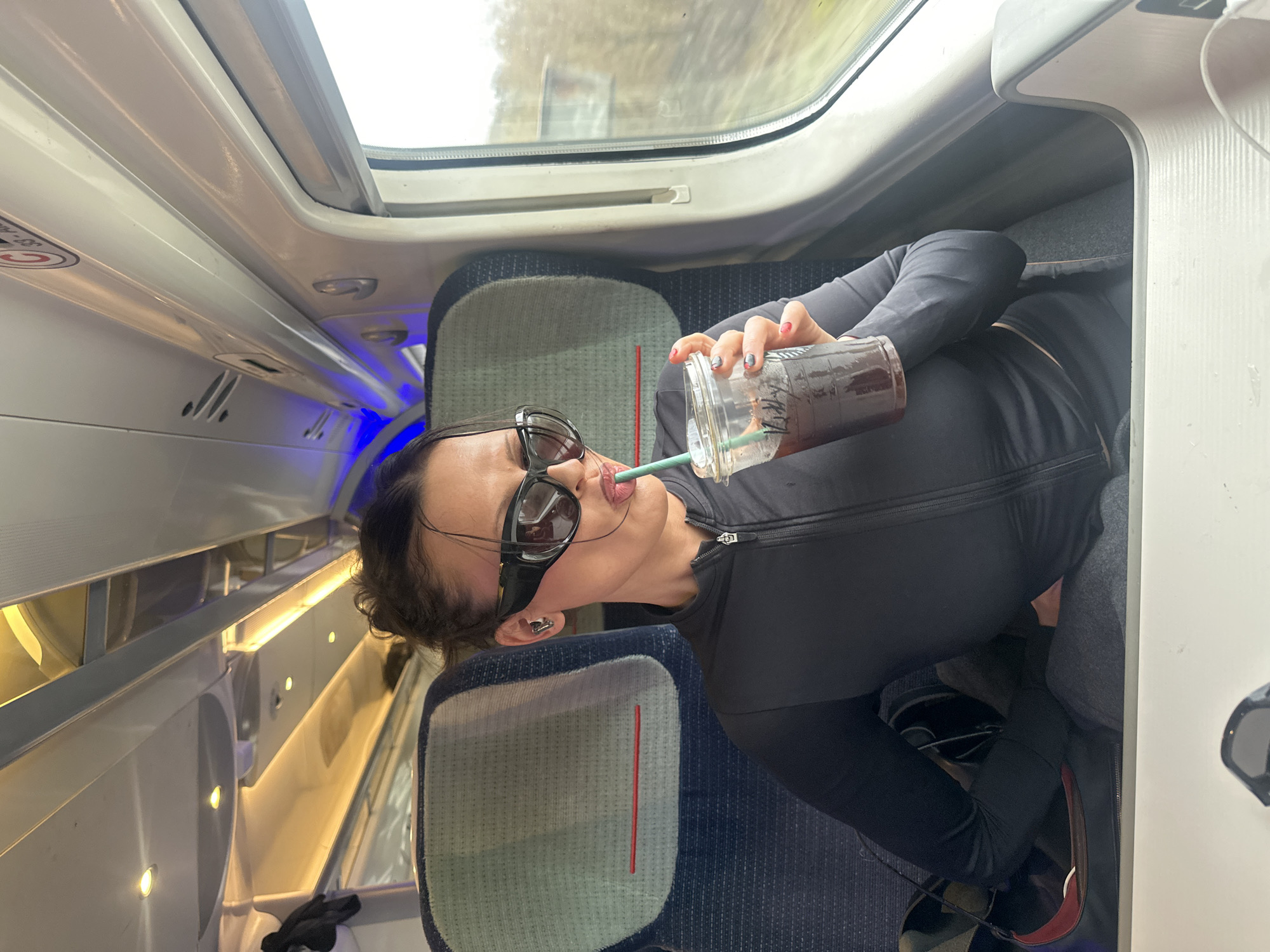 A female individual is seated aboard a Southeastern Class 395 "Javelin" train, operating on the High Speed 1 line within the United Kingdom, connecting to or from London St Pancras International. She wears a black long-sleeved zippered top, large dark sunglasses, and wireless earbuds in both ears. Her dark hair is styled back with some loose strands. She is actively drinking an iced beverage from a clear plastic cup, labeled "Kitty," through a green straw. Her right hand, featuring red and silver nail polish, holds the cup. The train's interior is visible, showing blue and grey upholstered seats, overhead luggage racks with recessed warm lighting, and additional blue accent lighting. A window on the right reveals a blurred green landscape, indicative of movement. A white table is positioned in front of the subject.