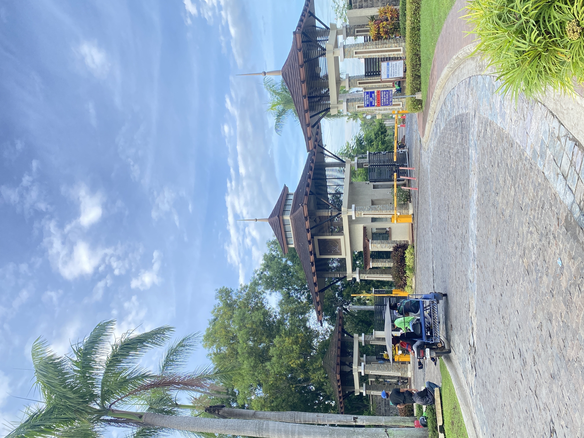 The image depicts the entrance to a residential development in Davao City, Philippines. The scene features two ornate guardhouses with distinctive brown, multi-tiered roofs and stone pillars. Yellow barriers, commonly used for vehicle control, are visible at the entrance. Several people are present: two individuals are seated on a grassy area to the left, while a three-wheeled vehicle, likely a tricycle or "kuliglig," carrying four passengers, is positioned on the cobblestone road. The environment is lush with palm trees and other greenery, and the sky is partly cloudy. Signage is visible near the guardhouses, indicating rules and information for residents and visitors. The overall impression is one of an orderly and well-maintained residential entrance in a tropical setting.