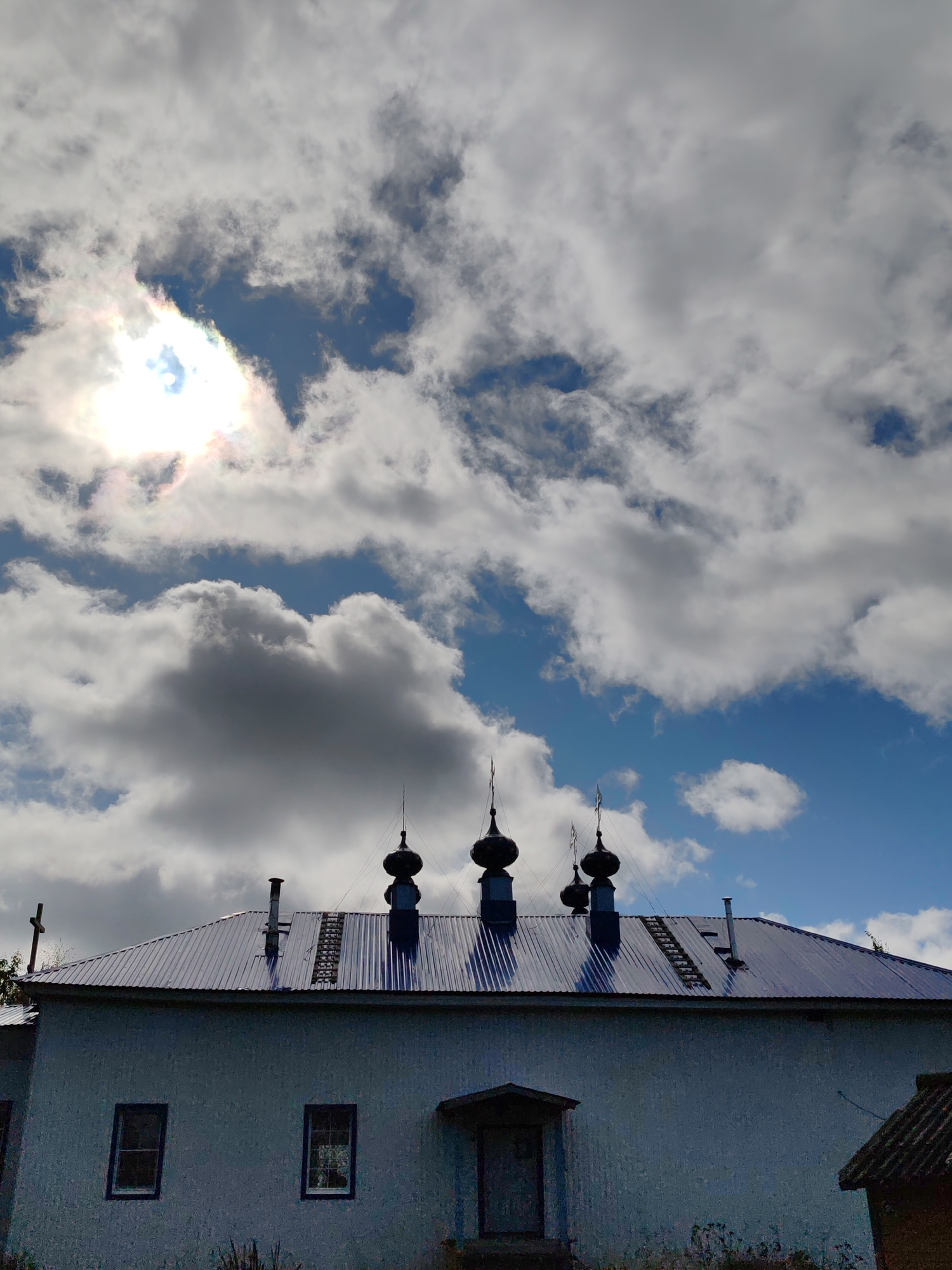 The image shows a view of a light blue building with a dark blue, corrugated metal roof, topped with several dark, onion-shaped domes, characteristic of Russian Orthodox churches. The building has two visible windows on its facade and a small entranceway. A ladder is affixed to the roof, leading up to the domes. In the background, a bright sun shines through dramatic, white and gray clouds against a blue sky. The scene appears to be set in a rural or semi-rural environment, possibly in the city of Chukhlem, Russia, given the architectural style. There are no people visible in the image.