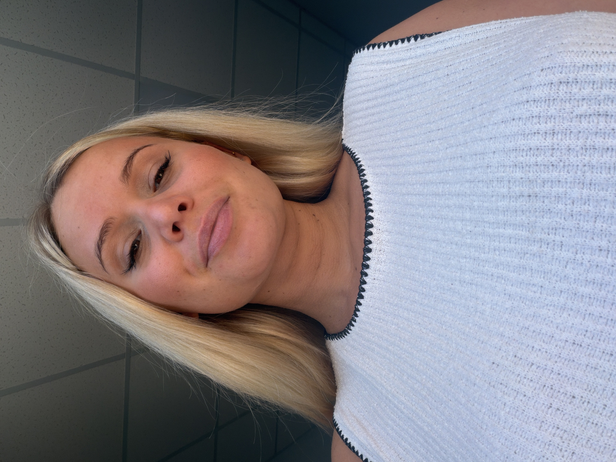A young Caucasian woman with blonde hair and fair skin is depicted in a close-up, angled shot. She is wearing a white, sleeveless top with a textured knit pattern and a black stitched trim around the neckline. Her makeup includes eyeliner and mascara, and she has freckles across her nose and cheeks. Her lips are slightly parted, and she is looking upwards and to her left. The background appears to be indoors, with a textured ceiling visible above her. The image is lit from above, casting soft shadows. The location is not identifiable as Sarapul, Russia.