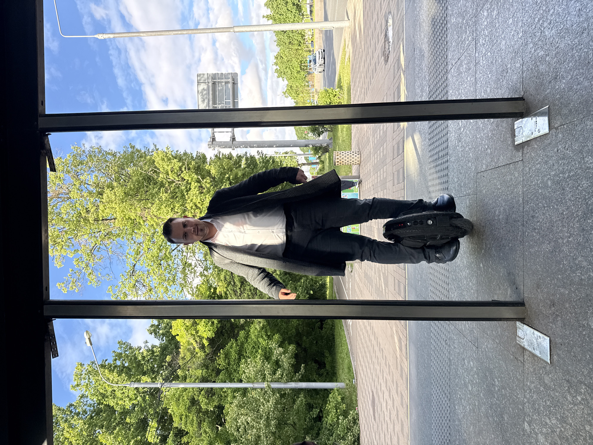 A man is standing on a one-wheeled electric scooter in front of a building. He is wearing dark pants, black shoes, a white shirt, and a gray jacket. The location appears to be Moscow, Russia, based on architectural style and street markings. The man is framed by a dark entryway, possibly a building entrance. Outside the entryway is a sidewalk, a green area with trees, a street, and street signs/lights. The weather is sunny with a blue sky and some clouds.