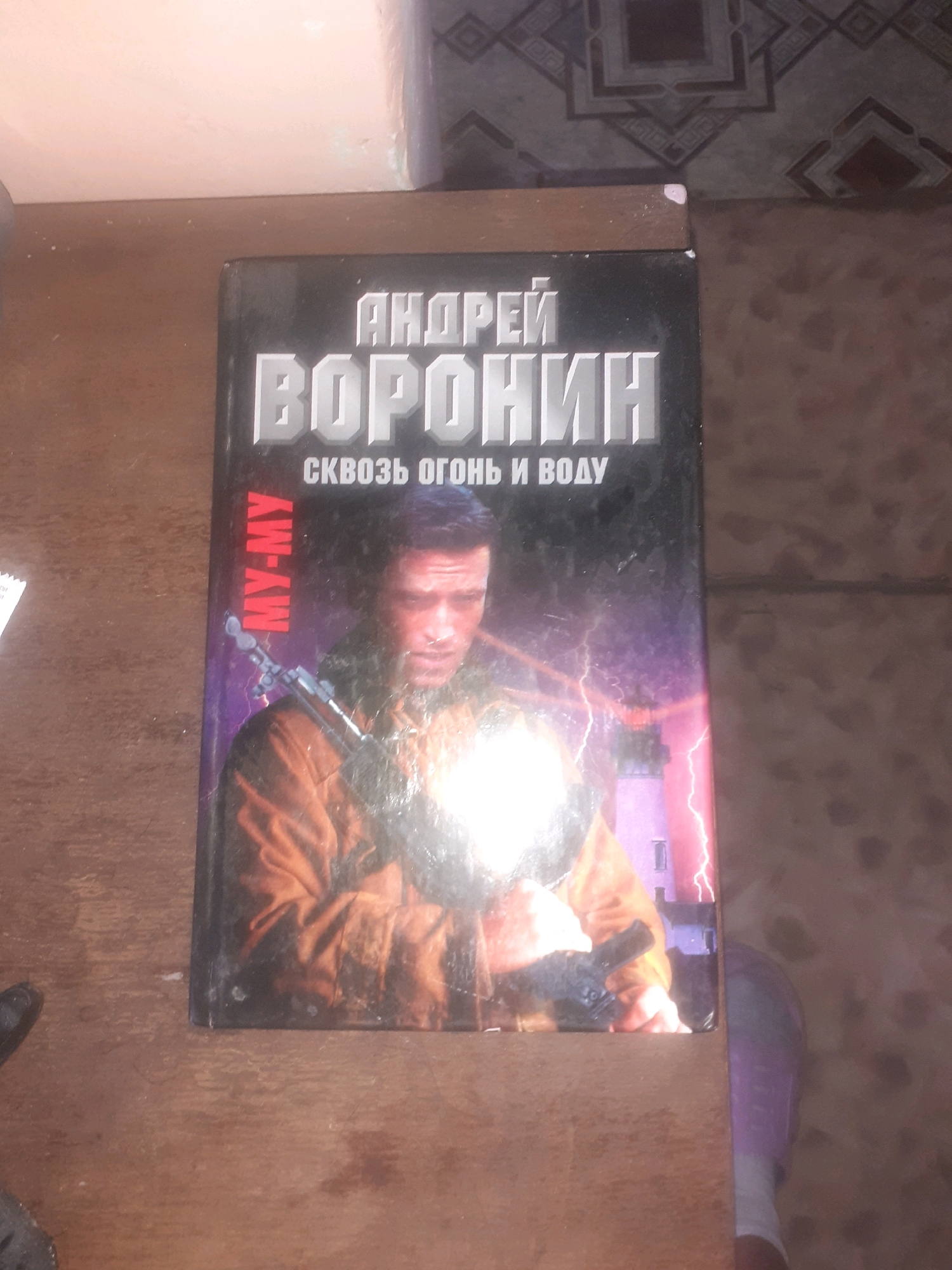 The image shows the cover of a book titled "Андрей Воронин сквозь огонь и воду". The cover features a close-up of a man holding a rifle, with a lighthouse and lightning in the background. The man appears to be in his 30s or 40s with short, dark hair and a serious expression. He is wearing a brown jacket. The text on the cover is in Russian. The book is placed on a wooden surface, likely a table. The lighting in the image is dim and there is a significant glare on the book cover, obscuring some details. The background appears to be a wall with a patterned wallpaper. The image does not contain any information that would allow for the identification of its location in Semey, Kazakhstan.
